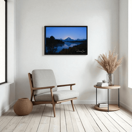 A modern living room features a light gray armchair, round side table with dried grasses, a woven basket, and "Sunrise at Oxbow Bend"—a framed Grand Teton art print by Rachna Jain from Mirabilia Boutique—on a white wall.