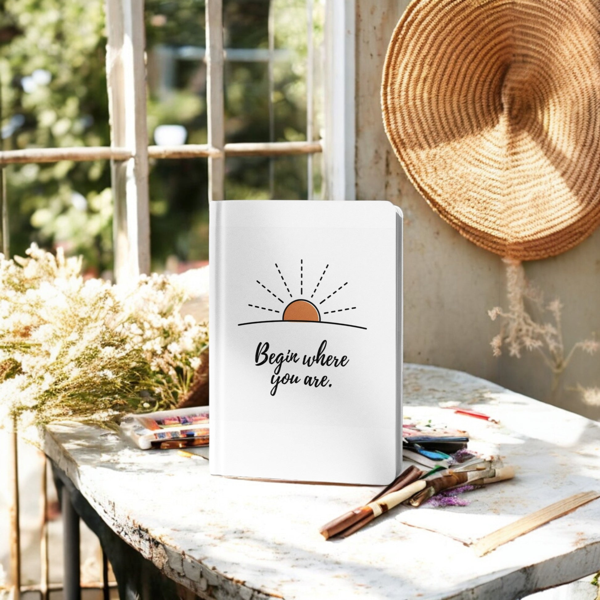 The Begin Where You Are Journal – Sunrise Quote Notebook by Mirabilia Boutique sits on a sunlit table with art supplies, dried flowers, and a woven hat nearby—perfect for daily writing amid greenery outside the window.
