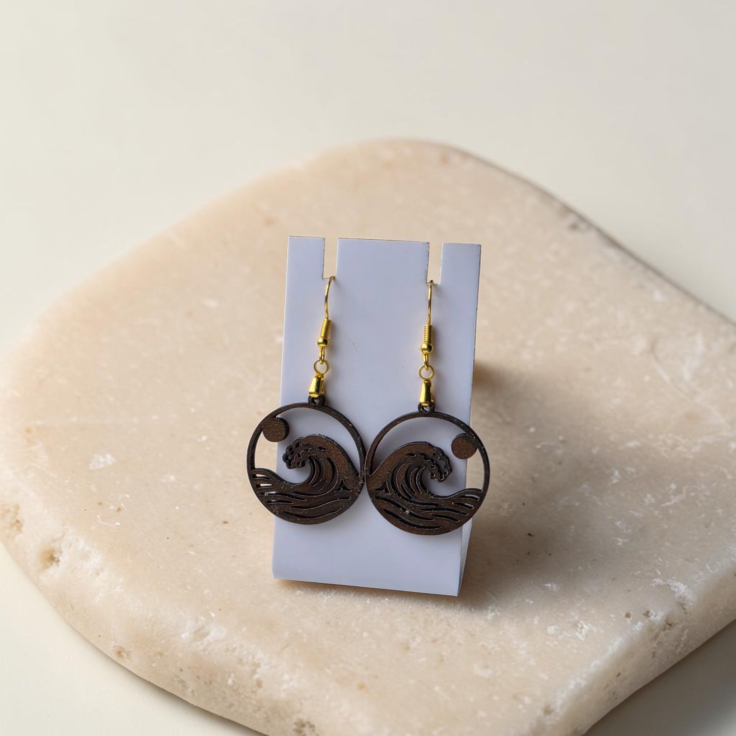 The Sunrise Wave Earrings by Mirabilia Boutique feature round, lasercut walnut wood with wave designs, displayed on a white card atop a beige stone—ideal coastal jewelry for ocean lovers.