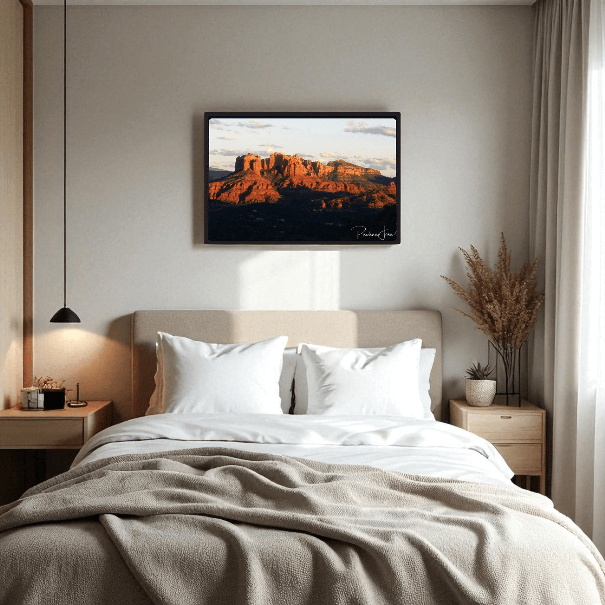 A cozy bedroom features a neatly made bed with two white pillows, beige blanket, wooden nightstands, a hanging lamp, dried plants in a vase, and "Sunset in Sedona" Southwest landscape framed art print by Rachna Jain from Mirabilia Boutique above the bed.