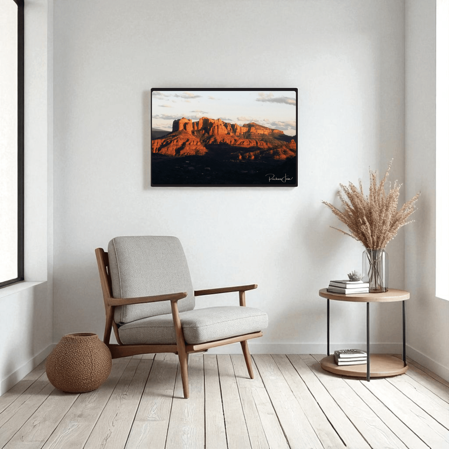 A modern, minimalist living room with a gray armchair, round side table holding a vase of dried grass, woven basket, and Mirabilia Boutique’s "Sunset in Sedona" framed art print by Rachna Jain on white walls with light wood floors.