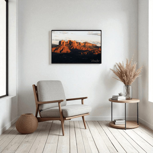 A modern, minimalist living room with a gray armchair, round side table holding a vase of dried grass, woven basket, and Mirabilia Boutique’s "Sunset in Sedona" framed art print by Rachna Jain on white walls with light wood floors.