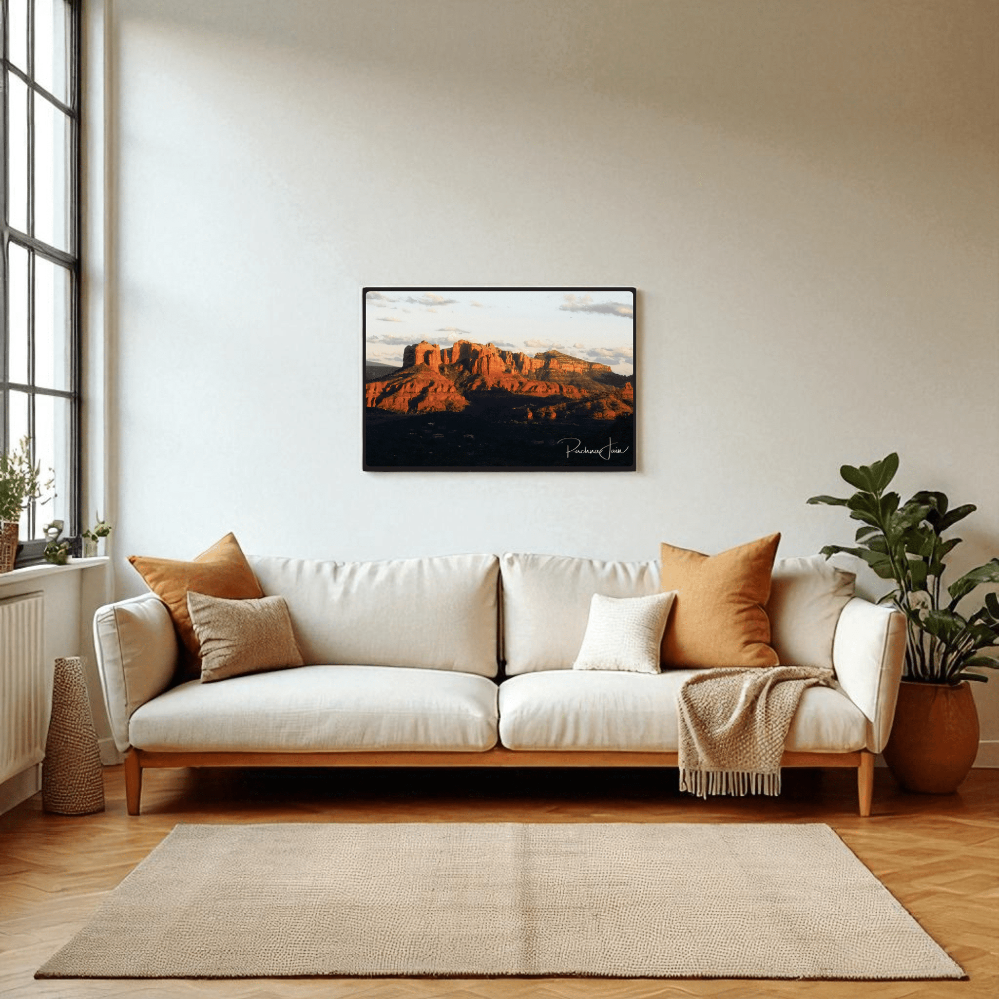 A bright living room features a white sofa, cozy pillows, potted plants, and a large window. Above the sofa hangs "Sunset in Sedona" framed art print by Rachna Jain from Mirabilia Boutique—a stunning southwest landscape.