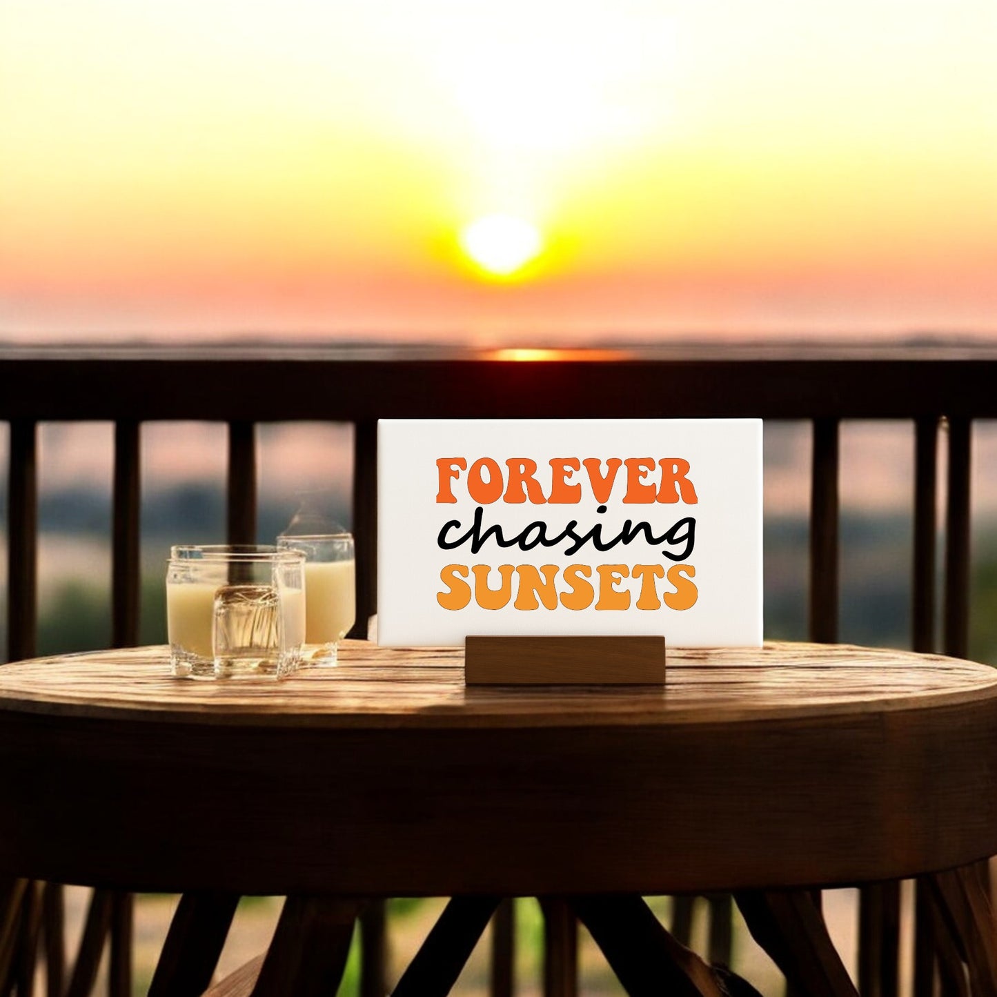 The Mirabilia Boutique Forever Chasing Sunsets Canvas – Retro Mini Sign sits on a wooden balcony table at sunset with two glasses, capturing the perfect summer vibe for inspirational decor or art display.