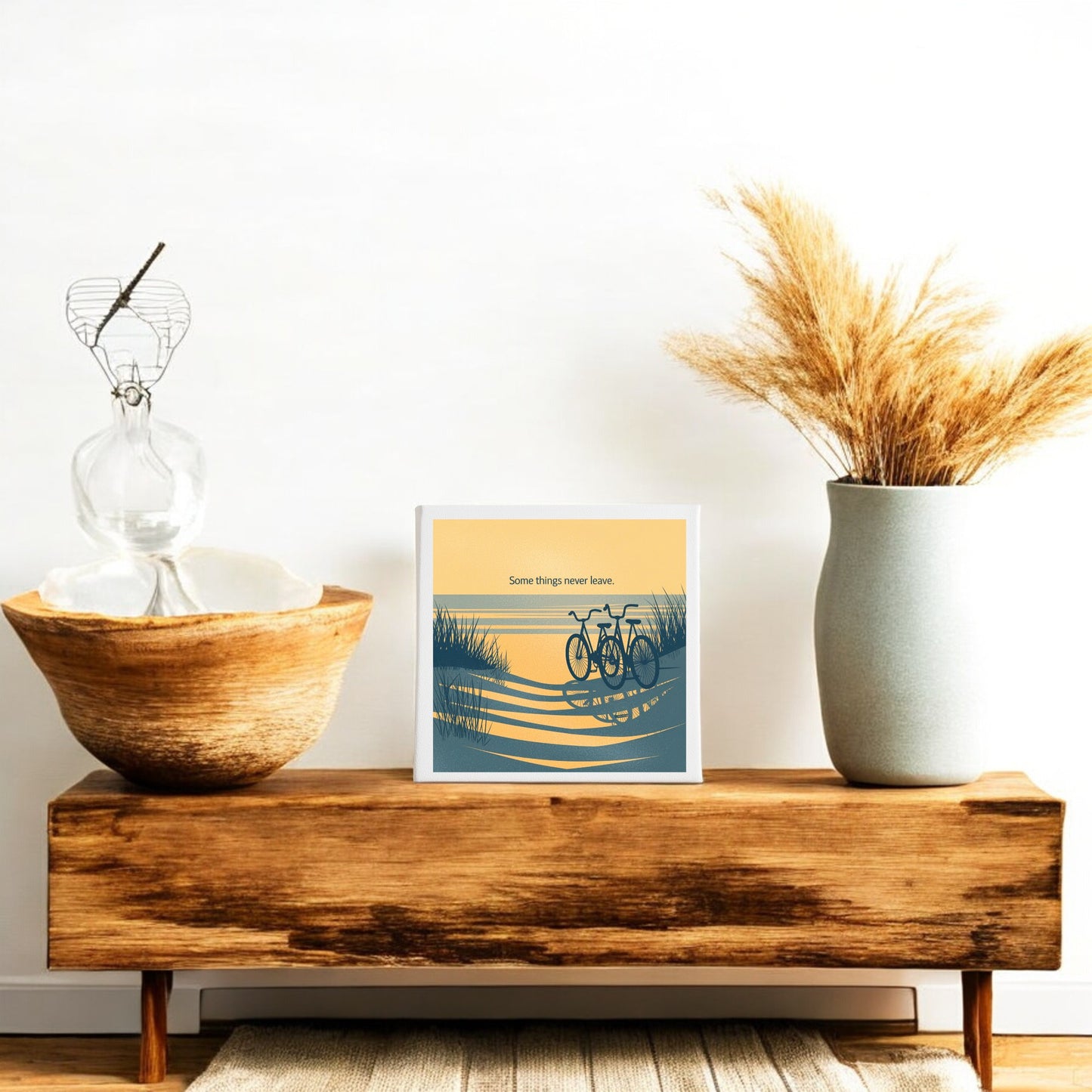 A wooden bench displays Mirabilia Boutique’s "Some Things Never Leave Canvas – Coastal Memory Art," a framed bicycle print, a large ceramic vase with dried grass, a glass sculpture, and a wooden bowl for cozy minimalist decor against a white wall.