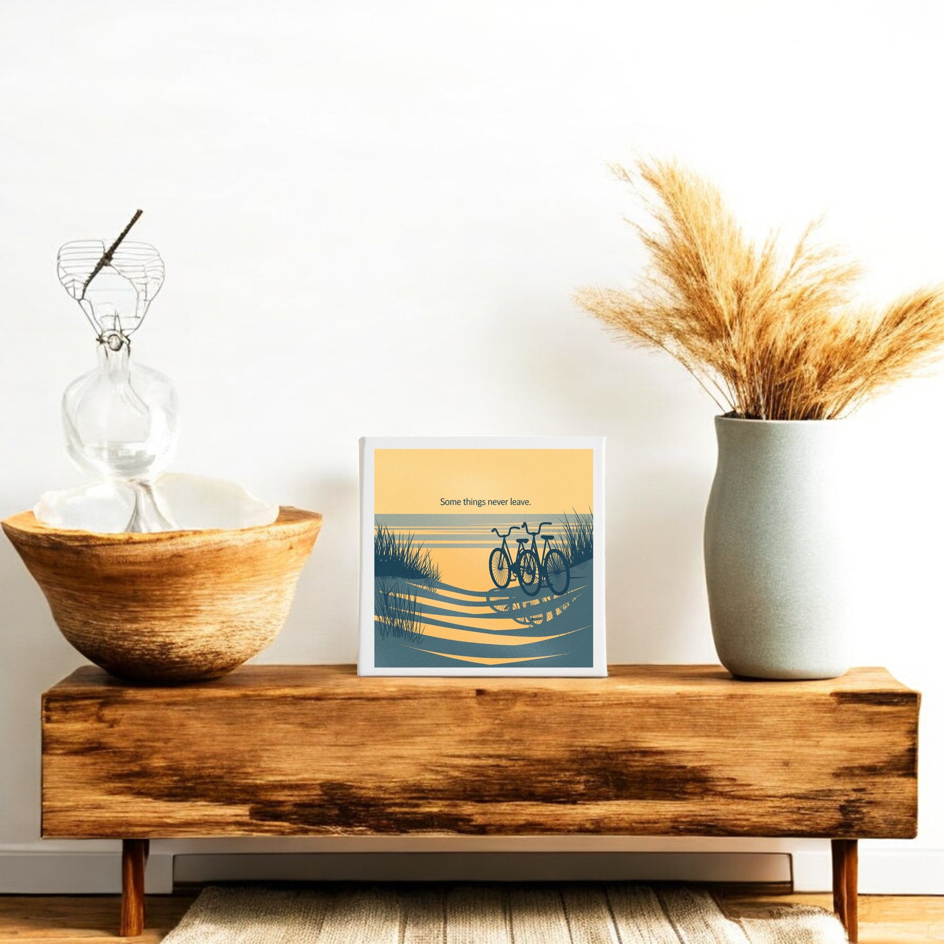 A wooden bench displays Mirabilia Boutique’s "Some Things Never Leave Canvas – Coastal Memory Art," a framed bicycle print, a large ceramic vase with dried grass, a glass sculpture, and a wooden bowl for cozy minimalist decor against a white wall.