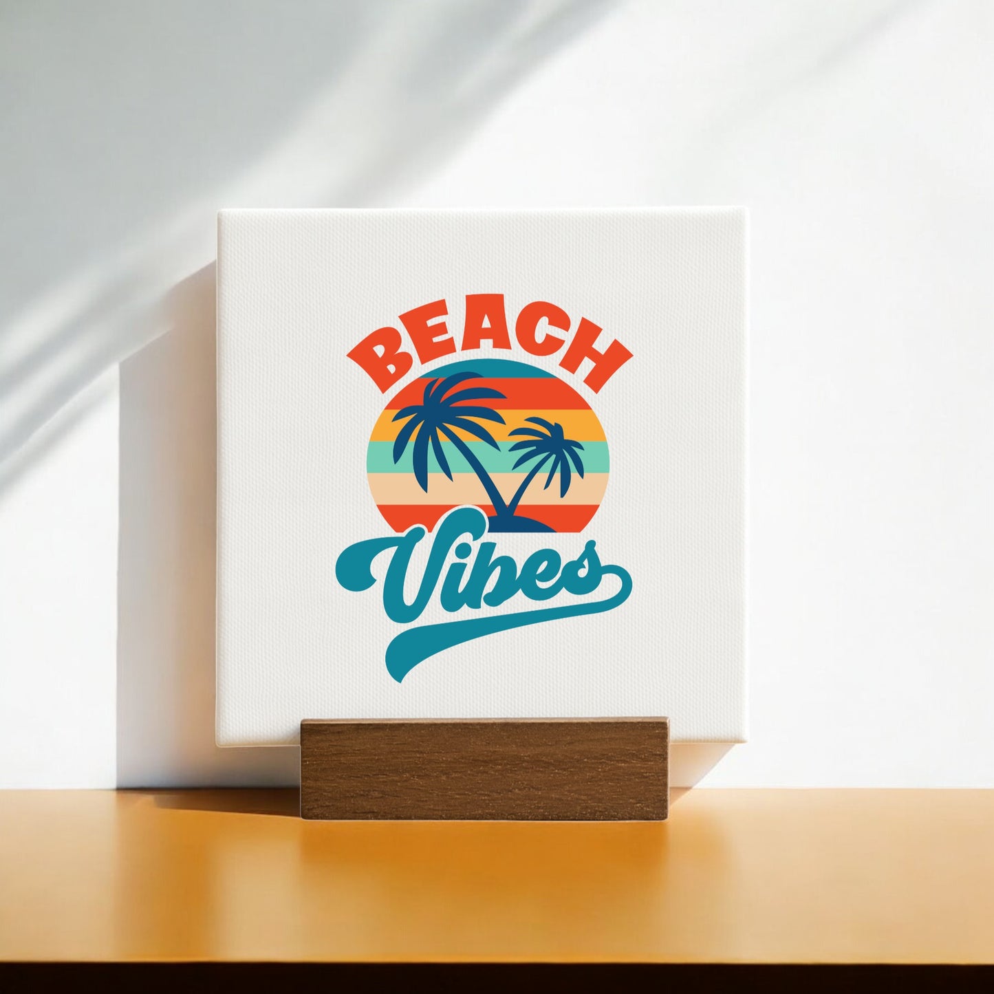 The Beach Vibes Sunset Canvas – Bold Tropical Mini Sign by Mirabilia Boutique features a retro sunset, palm trees, and vibrant lettering on a wooden stand. Sunlight and soft shadows enhance this charming coastal decor accent.