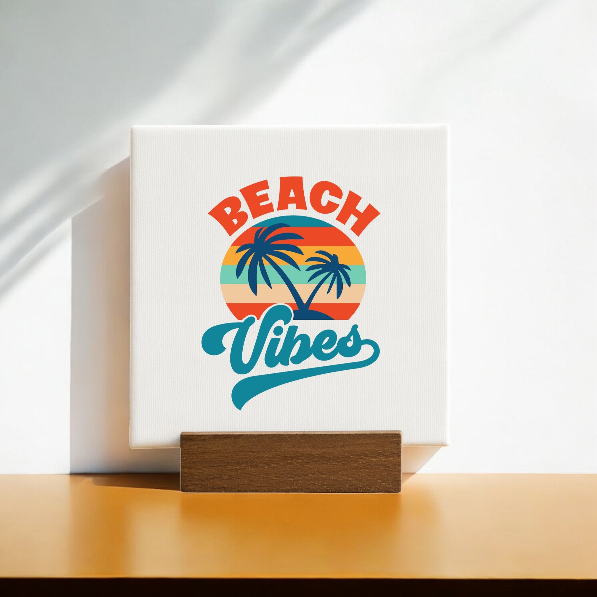 The Beach Vibes Sunset Canvas – Bold Tropical Mini Sign by Mirabilia Boutique features a retro sunset, palm trees, and vibrant lettering on a wooden stand. Sunlight and soft shadows enhance this charming coastal decor accent.