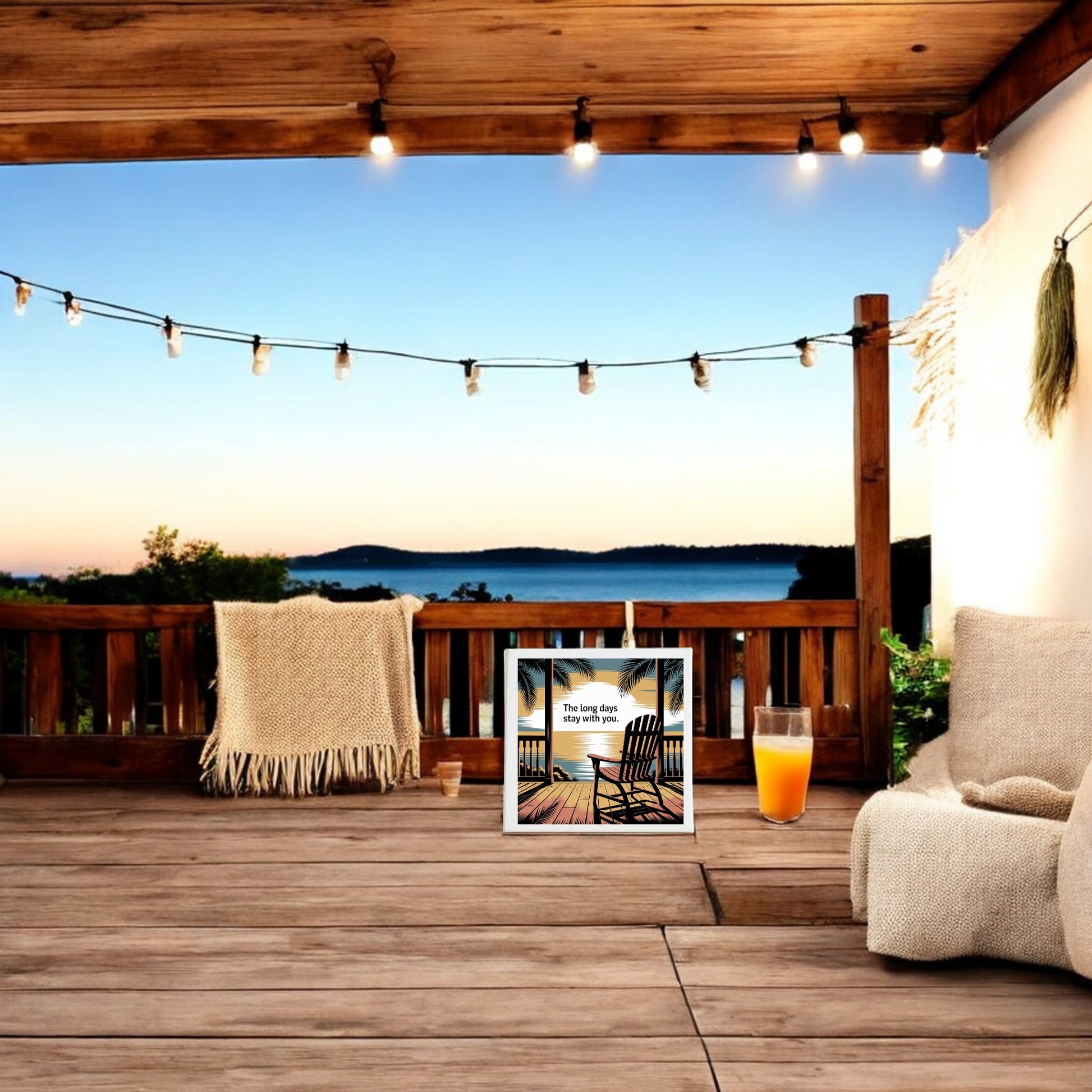 A cozy wooden patio with string lights overlooks a calm lake at sunset. The Long Days Canvas – Sunset Memory Porch Art by Mirabilia Boutique and a blanket-covered chair create a relaxed, inviting atmosphere with peaceful wall decor.