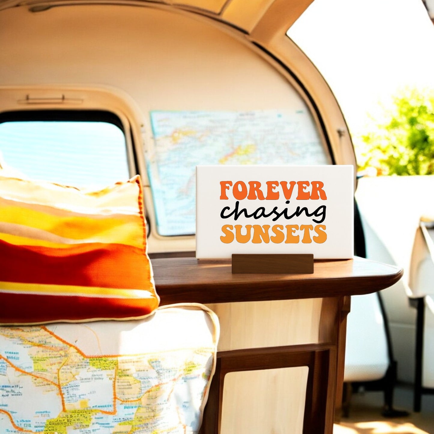 The Mirabilia Boutique “Forever Chasing Sunsets Canvas – Retro Mini Sign” sits on a camper van table with striped pillows, a map, and sunlight—a perfect sunset chaser decor accent.
