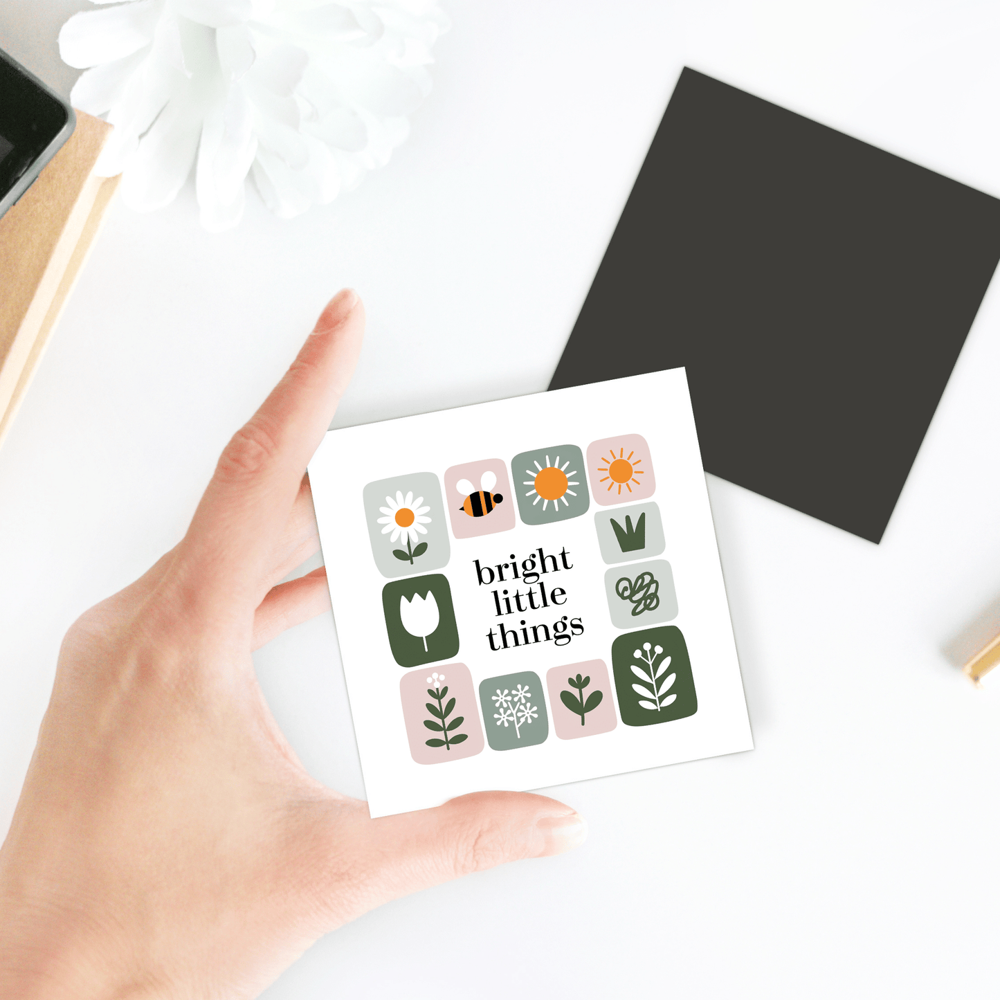 A hand holds the Bright Little Things Floral Grid Magnet by Mirabilia Boutique, featuring flower and bee illustrations. In the background, a black square, white flower, and notebooks add to the cheerful decor on the white surface.