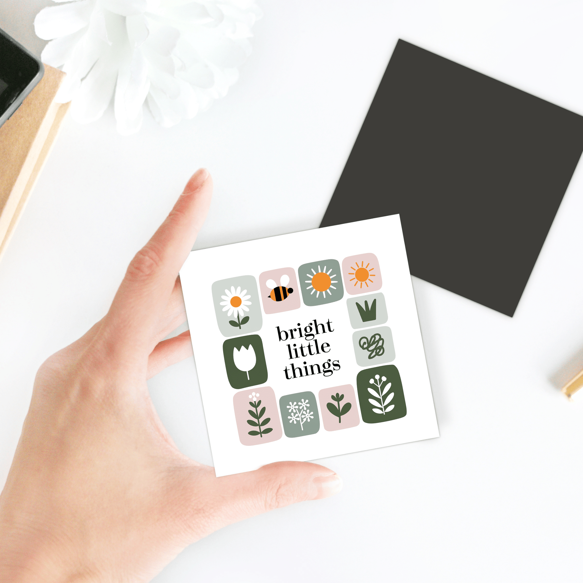 A hand holds the Bright Little Things Floral Grid Magnet by Mirabilia Boutique, featuring flower and bee illustrations. In the background, a black square, white flower, and notebooks add to the cheerful decor on the white surface.