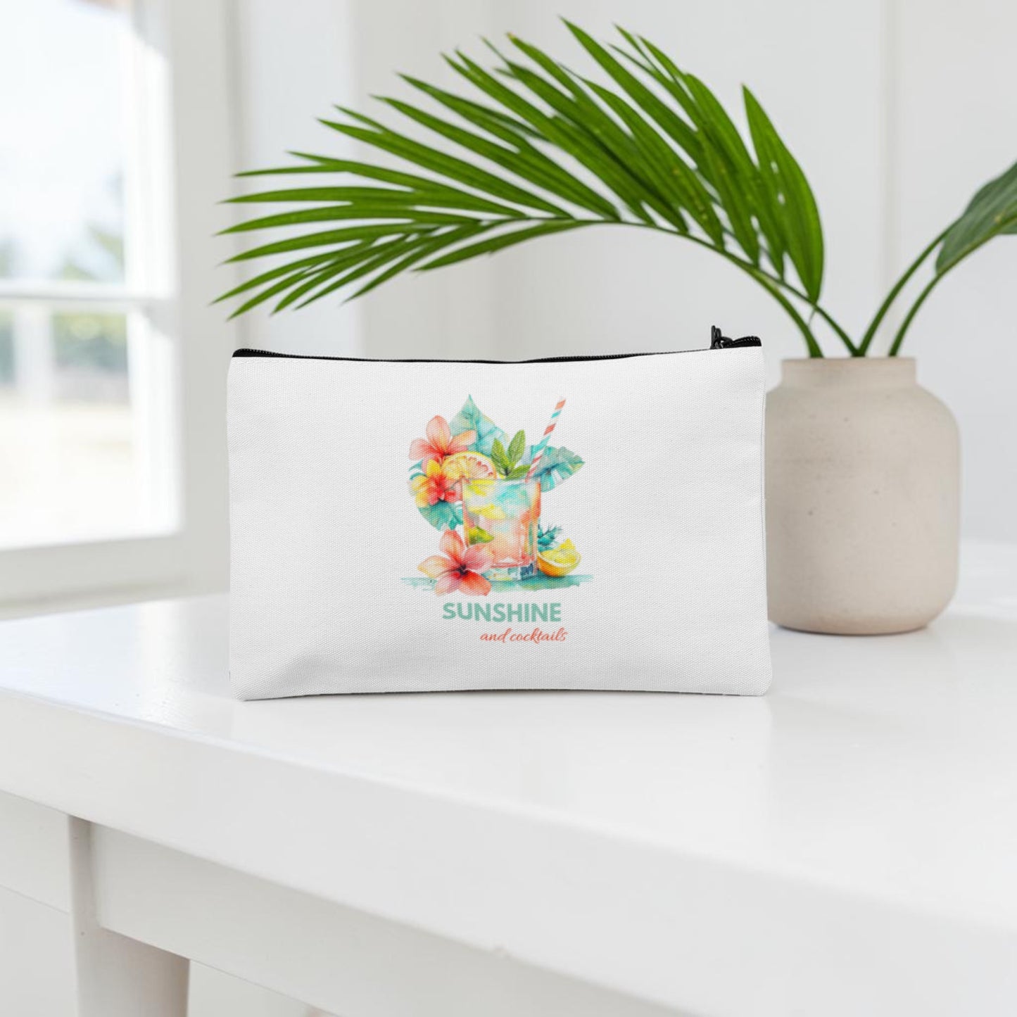 A "Sunshine and Cocktails Makeup Bag" by Mirabilia Boutique, featuring a white zippered design with a colorful tropical drink and flowers and the label "SUNSHINE and cocktails," sits on a white table next to a beige vase holding a green palm leaf.