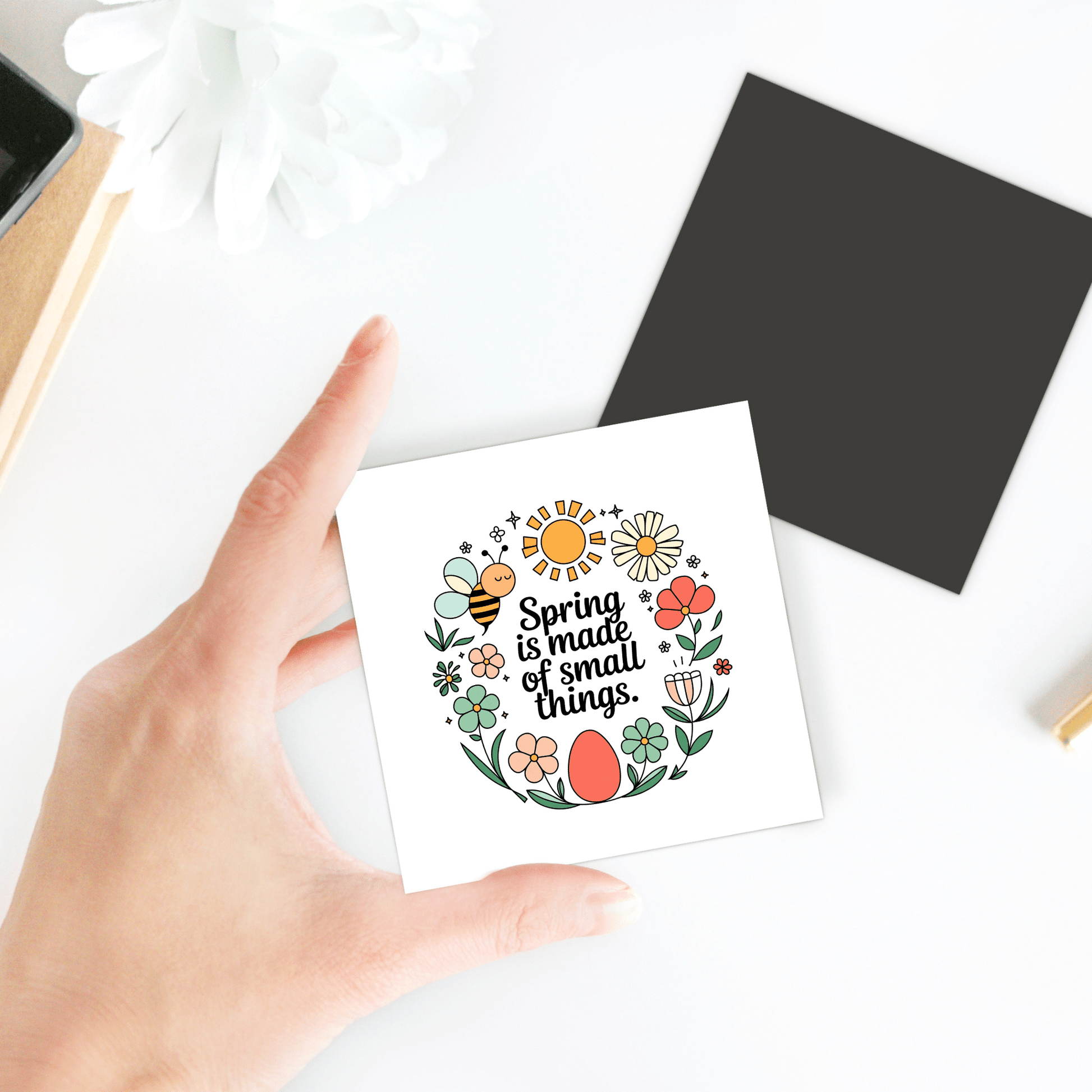 A hand holds the Mirabilia Boutique Spring Is Made of Small Things Floral Magnet, featuring flowers, an egg, and the phrase "Spring is made of small things," beside a black square card and white flower—ideal as an Easter gift.