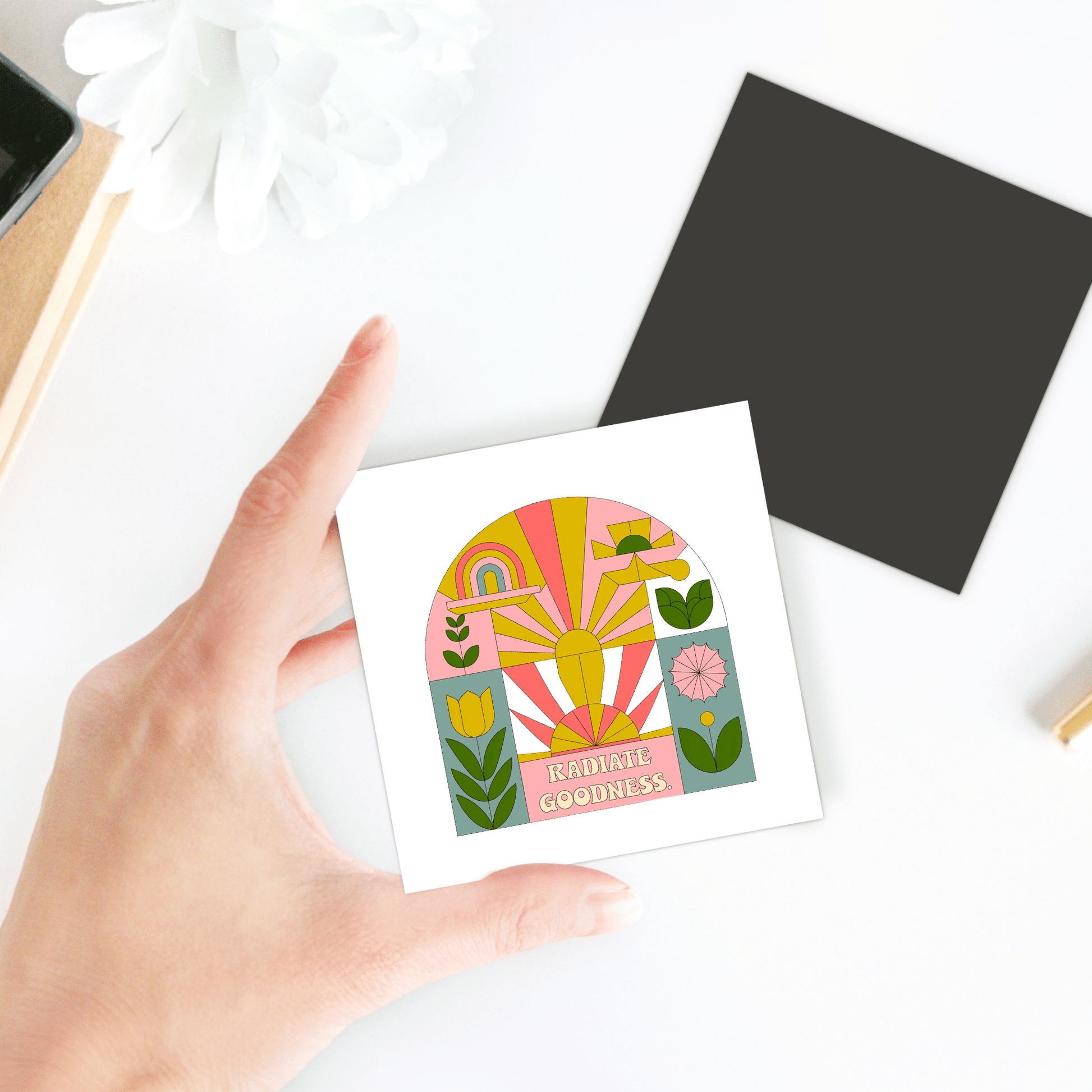 A hand holds the Radiate Goodness Colorful Floral Magnet by Mirabilia Boutique, featuring sunburst, rainbow, and flower designs. The magnet is on a white surface beside a dark square and a white flower.