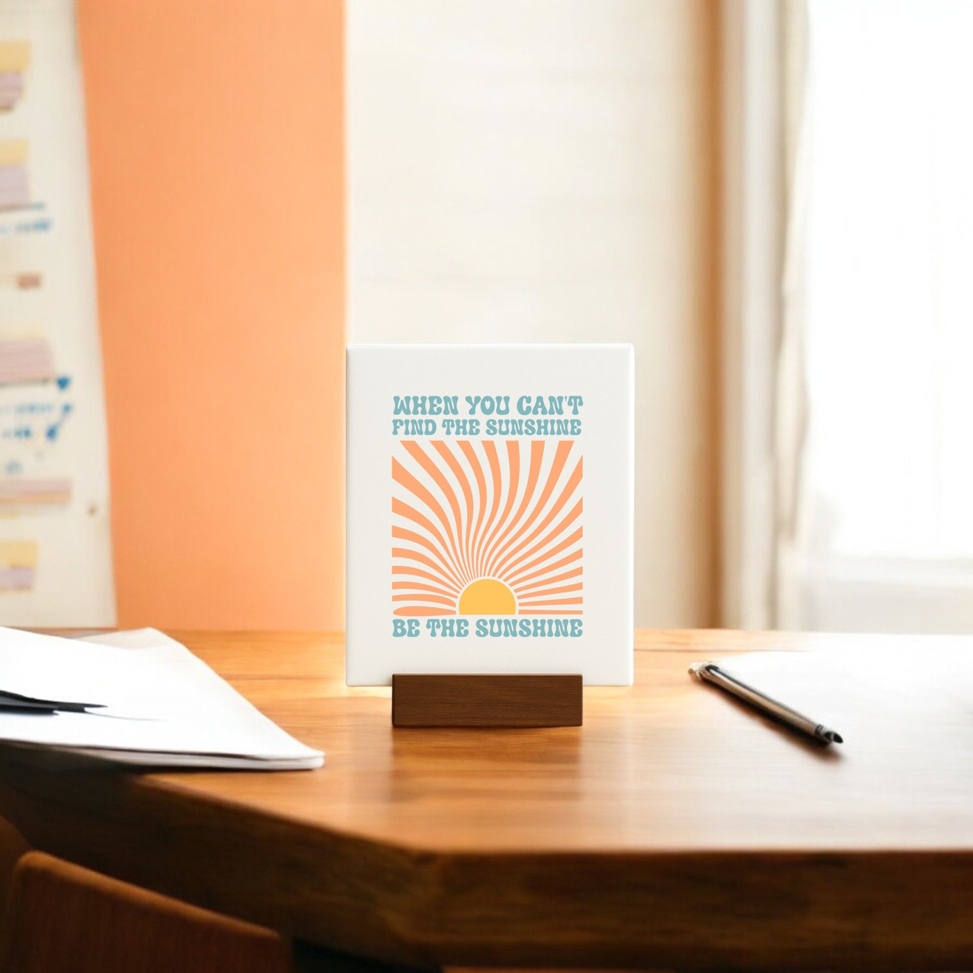 The "Be the Sunshine Mini Canvas – Retro Sunburst Sign" by Mirabilia Boutique sits on a wooden stand, featuring an uplifting quote and orange sun illustration—an inspiring desk accent beside a pen and papers.