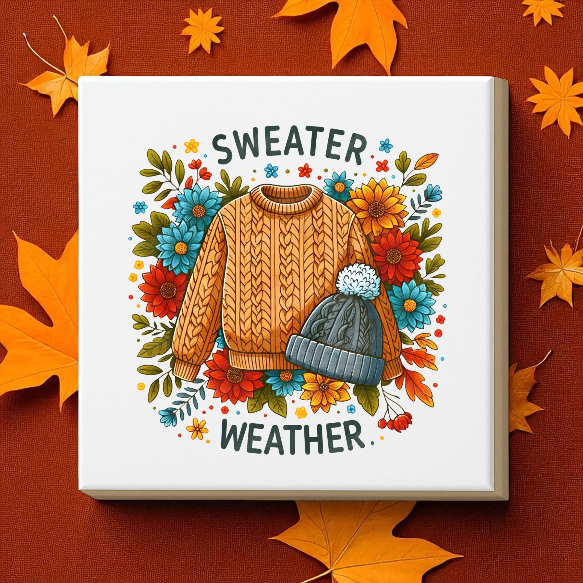 The Mirabilia Boutique "Sweater Weather Canvas" (8x8) features a cozy orange sweater and gray pom-pom hat surrounded by autumn flowers and leaves—perfect fall wall art to warm up your home décor.