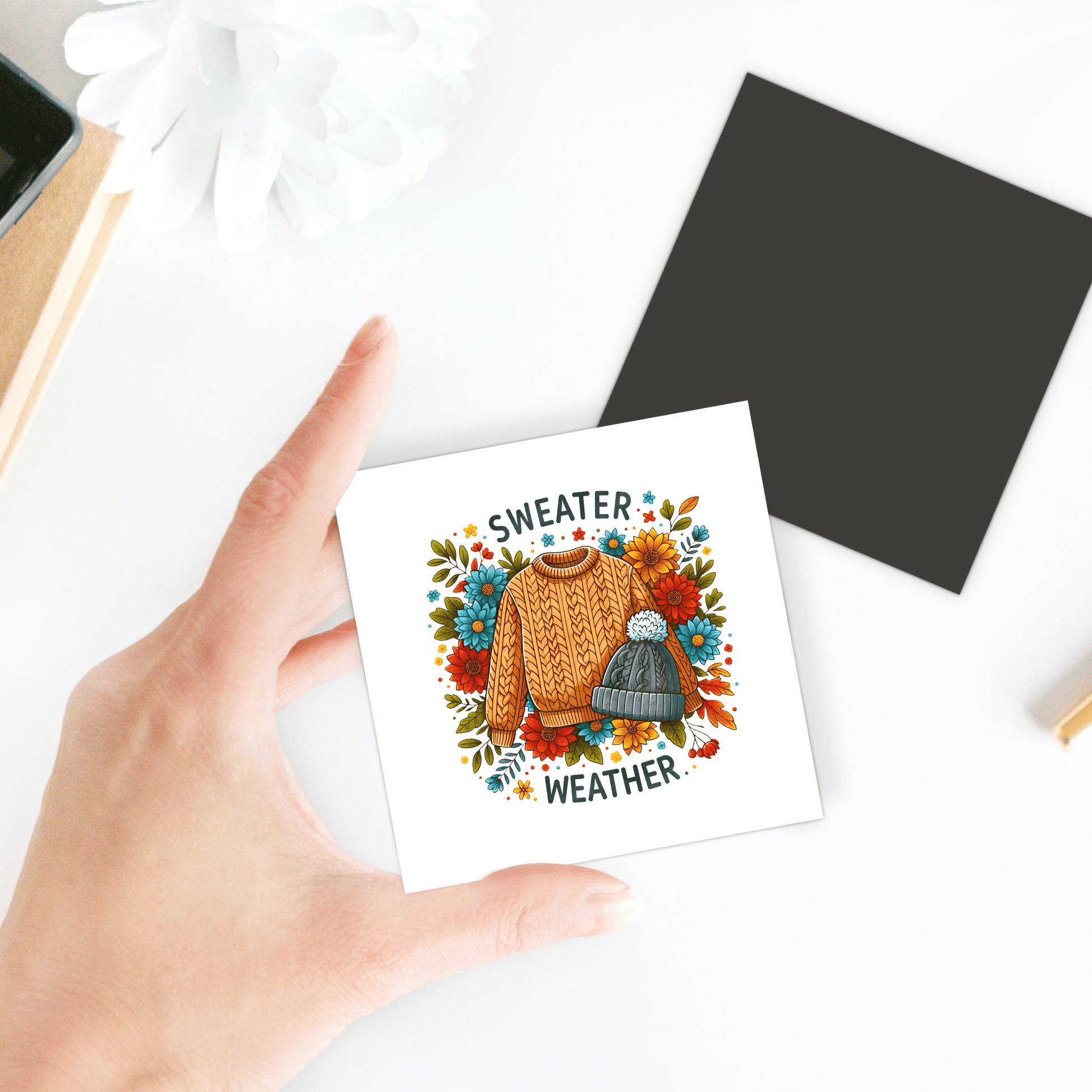 A hand holds a "Sweater Weather" magnet by Mirabilia Boutique, featuring cozy sweater art with a hat and autumn flowers—perfect fall fridge decor or an autumn gift.