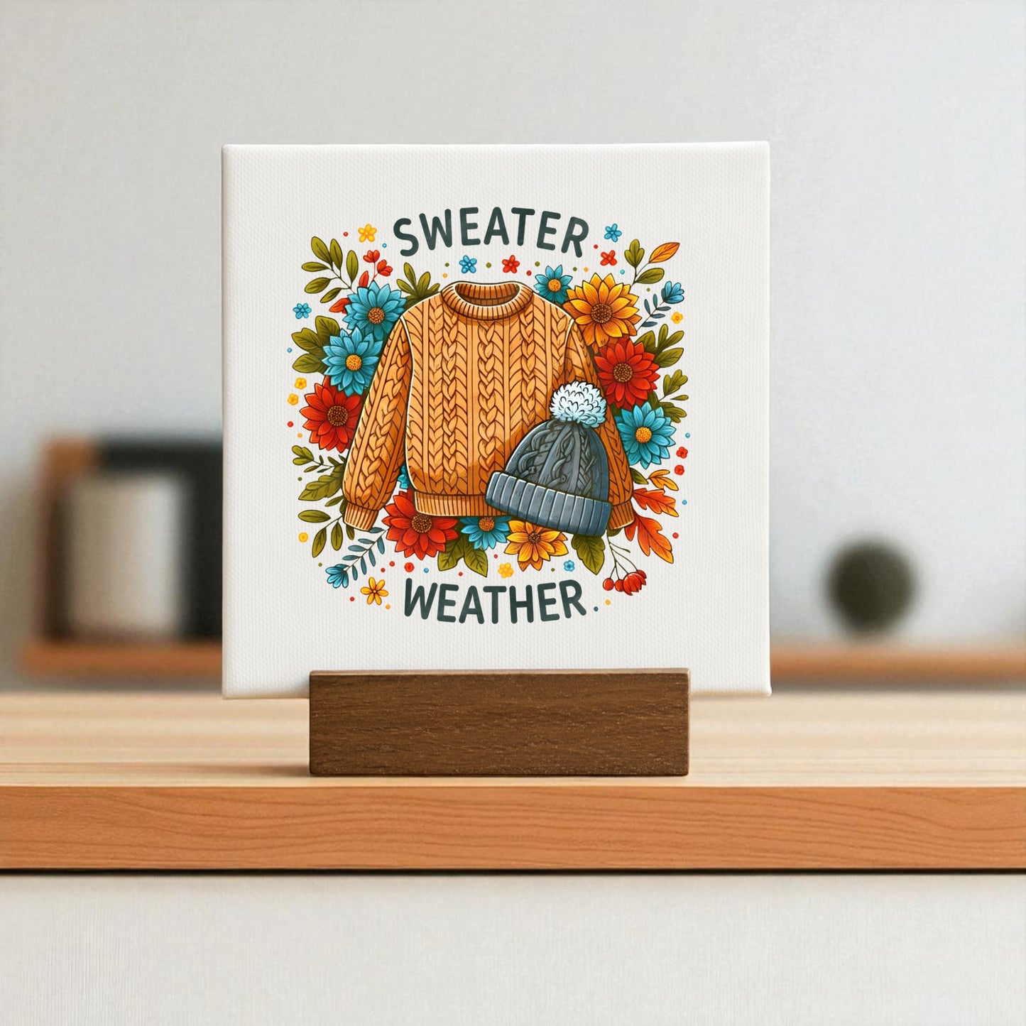 The Sweater Weather Mini Sign by Mirabilia Boutique features an orange sweater and gray knit hat on canvas, accented with autumn florals—making this cozy cable knit canvas a perfect fall home decor accent.