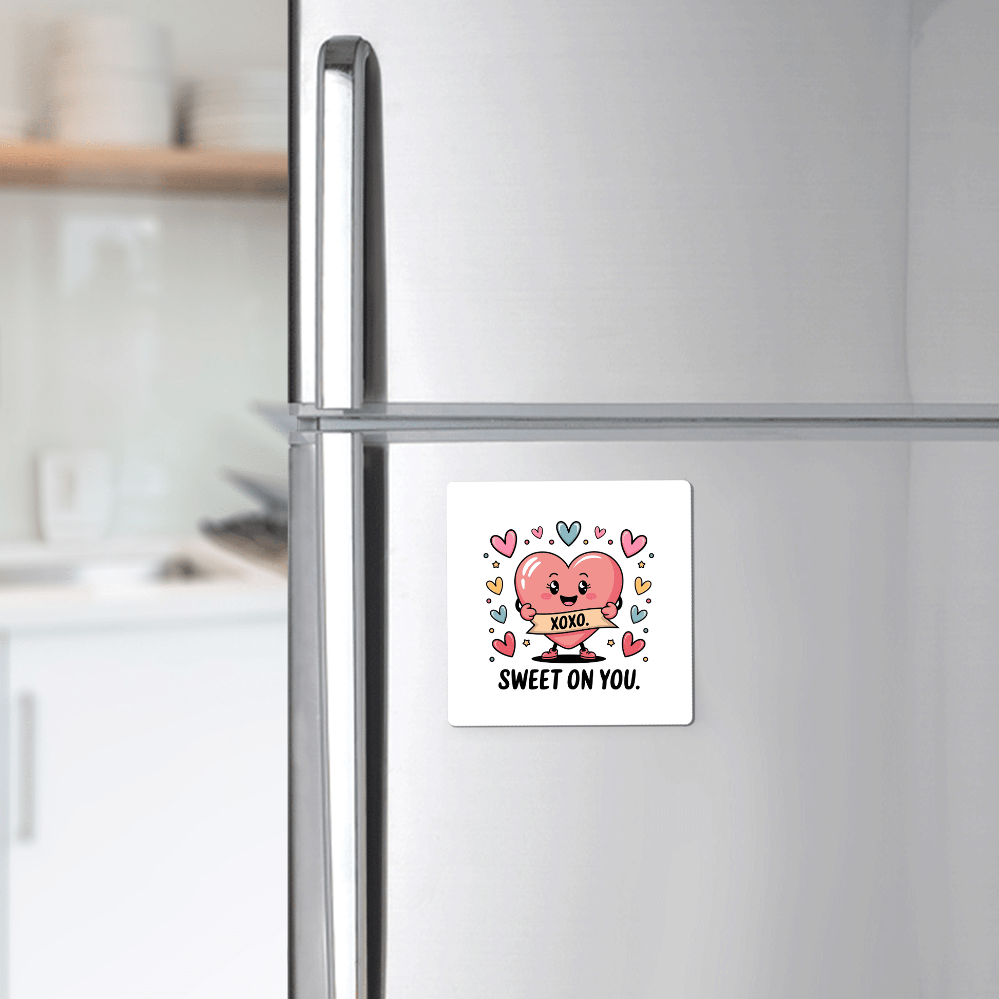 A silver refrigerator displays the Mirabilia Boutique "Sweet on You Valentine Magnet," featuring a pink heart cartoon, colorful hearts, and "SWEET ON YOU" text—adding romantic flair to the kitchen decor. The kitchen is seen in the background.