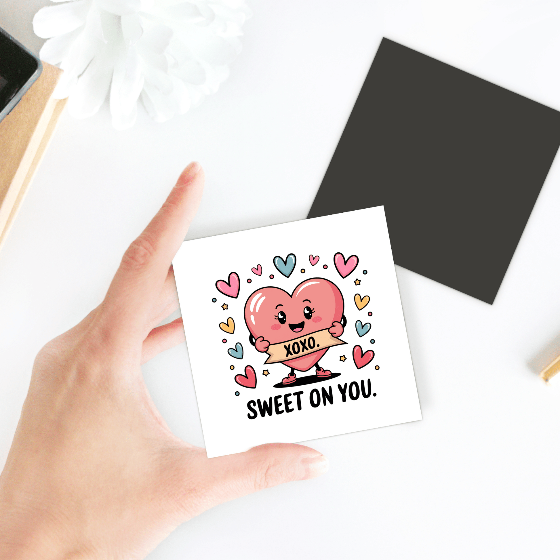 A hand holds the Sweet on You Valentine Magnet by Mirabilia Boutique—a square card with a pink heart character, colorful hearts, and an "XOXO" banner. Perfect as a romantic kitchen decor or love note. Flower and blank card accompany it on a white surface.