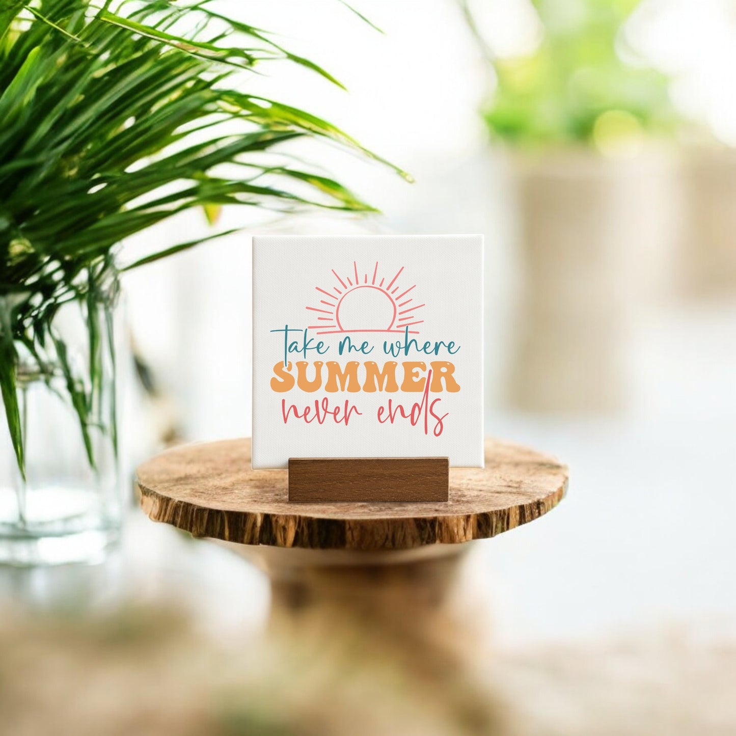 The Take Me Where Summer Never Ends Mini Canvas by Mirabilia Boutique features a sun illustration and sits on a wooden stand—ideal summer decor or beach house accent, beautifully displayed among greenery in soft natural light.