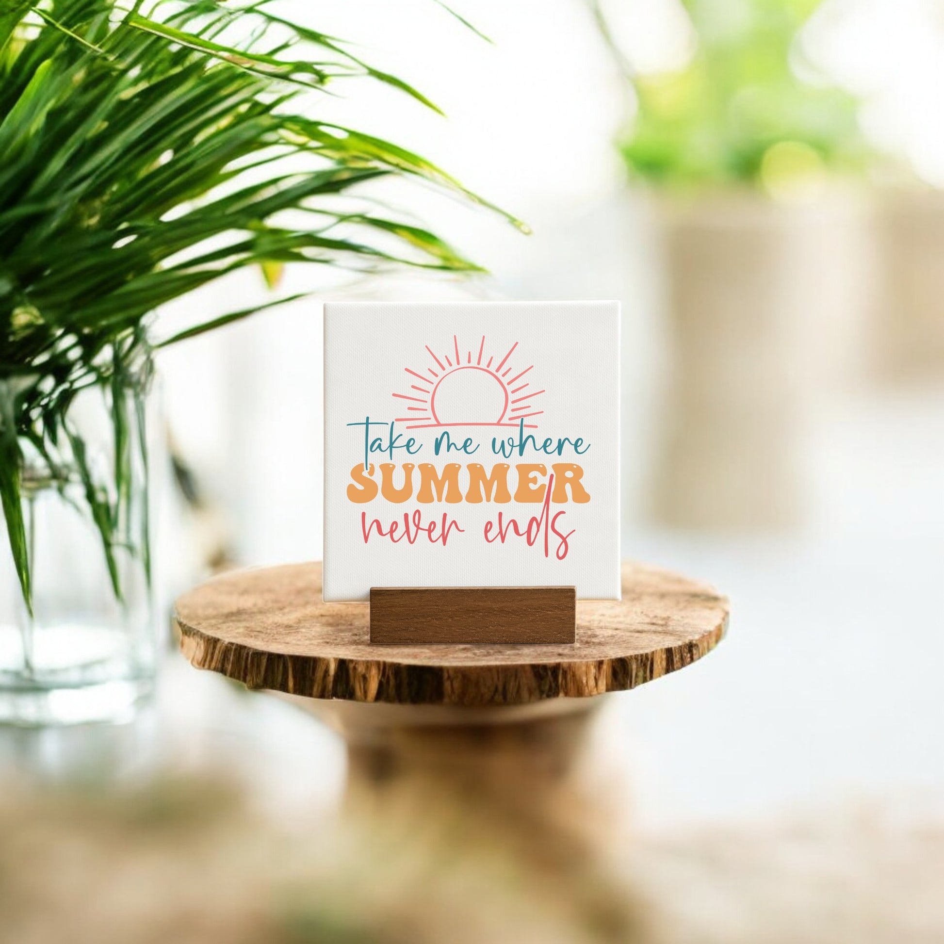 The Take Me Where Summer Never Ends Mini Canvas by Mirabilia Boutique features a sun illustration and sits on a wooden stand—ideal summer decor or beach house accent, beautifully displayed among greenery in soft natural light.