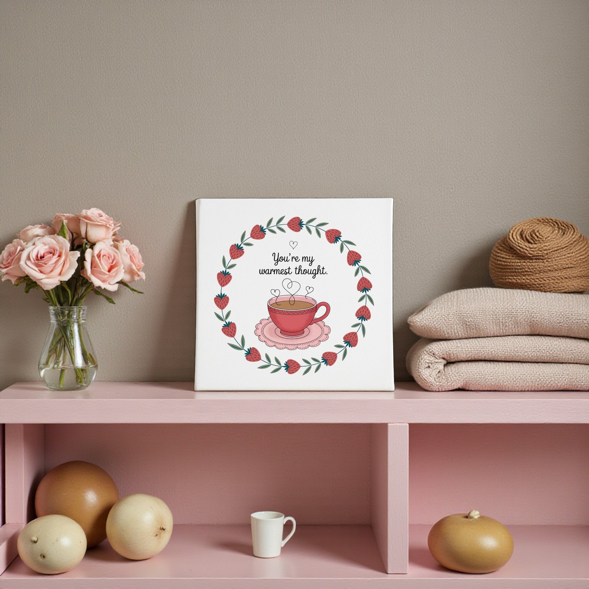 A pink shelf holds pink roses, beige blankets, twine balls, and melons. At the center is Mirabilia Boutique’s Warmest Thought Tea-Inspired Canvas Art—a lovely tea lover gift or sweet Valentine’s decor with its charming teacup design.