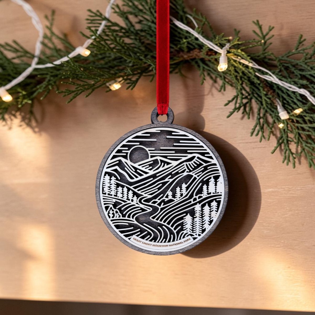 The Great Smoky Mountains National Park Line Art Ornament by Mirabilia Boutique features an oval design with a mountain and forest scene, elegantly hanging from a red ribbon. Accented by pine garland and string lights in the background, this ornament brings a festive touch—making it a delightful National Park keepsake.