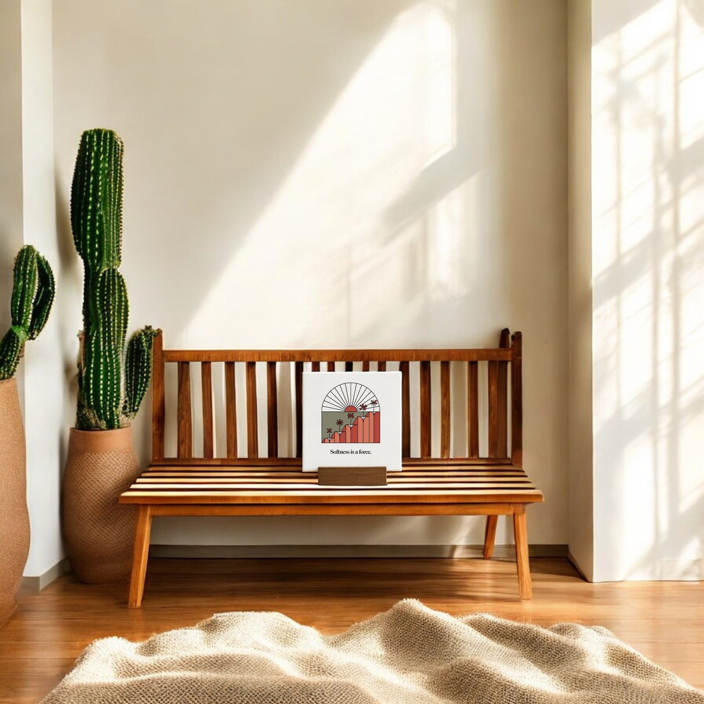 A wooden bench in a sunlit room displays the "Softness Is a Force" mini canvas sign by Mirabilia Boutique. Two tall cacti stand nearby, soft shadows fall, and the scene creates a cozy spot for your motivational desk decor.