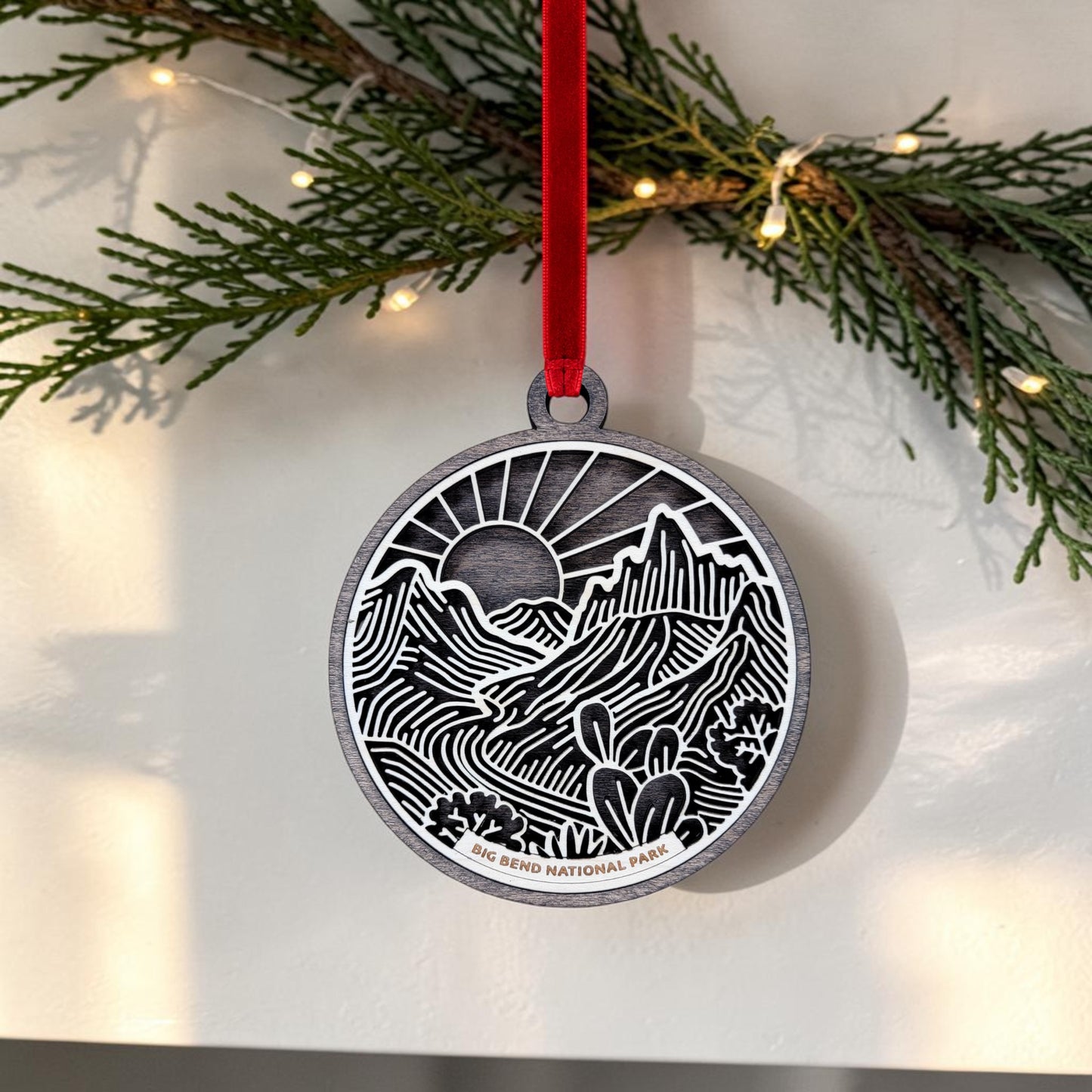 The Mirabilia Boutique Big Bend National Park Line Art Ornament is a round wooden keepsake, featuring a mountain and cactus landscape in Southwest line art style and suspended from a red ribbon. Against a backdrop of evergreen branches and string lights, this ornament displays the words "Big Bend National Park.