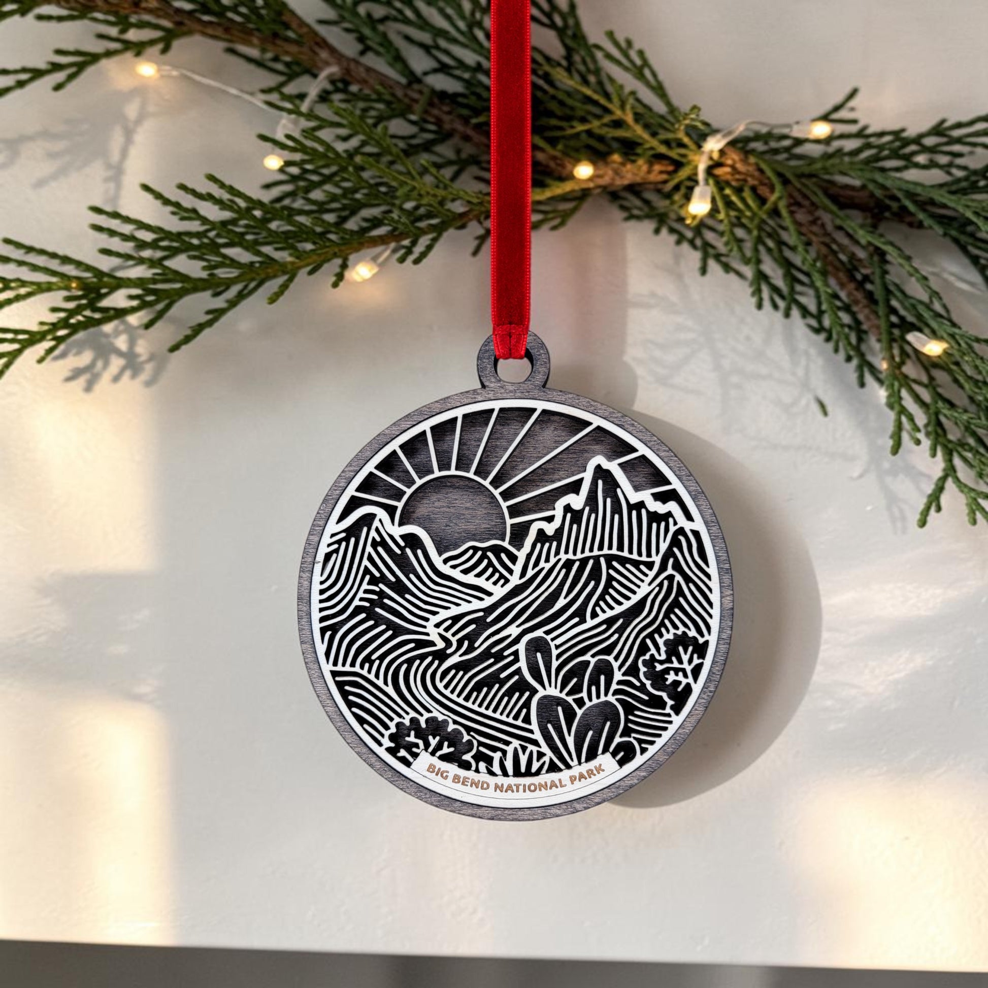 The Mirabilia Boutique Big Bend National Park Line Art Ornament is a round wooden keepsake, featuring a mountain and cactus landscape in Southwest line art style and suspended from a red ribbon. Against a backdrop of evergreen branches and string lights, this ornament displays the words "Big Bend National Park.