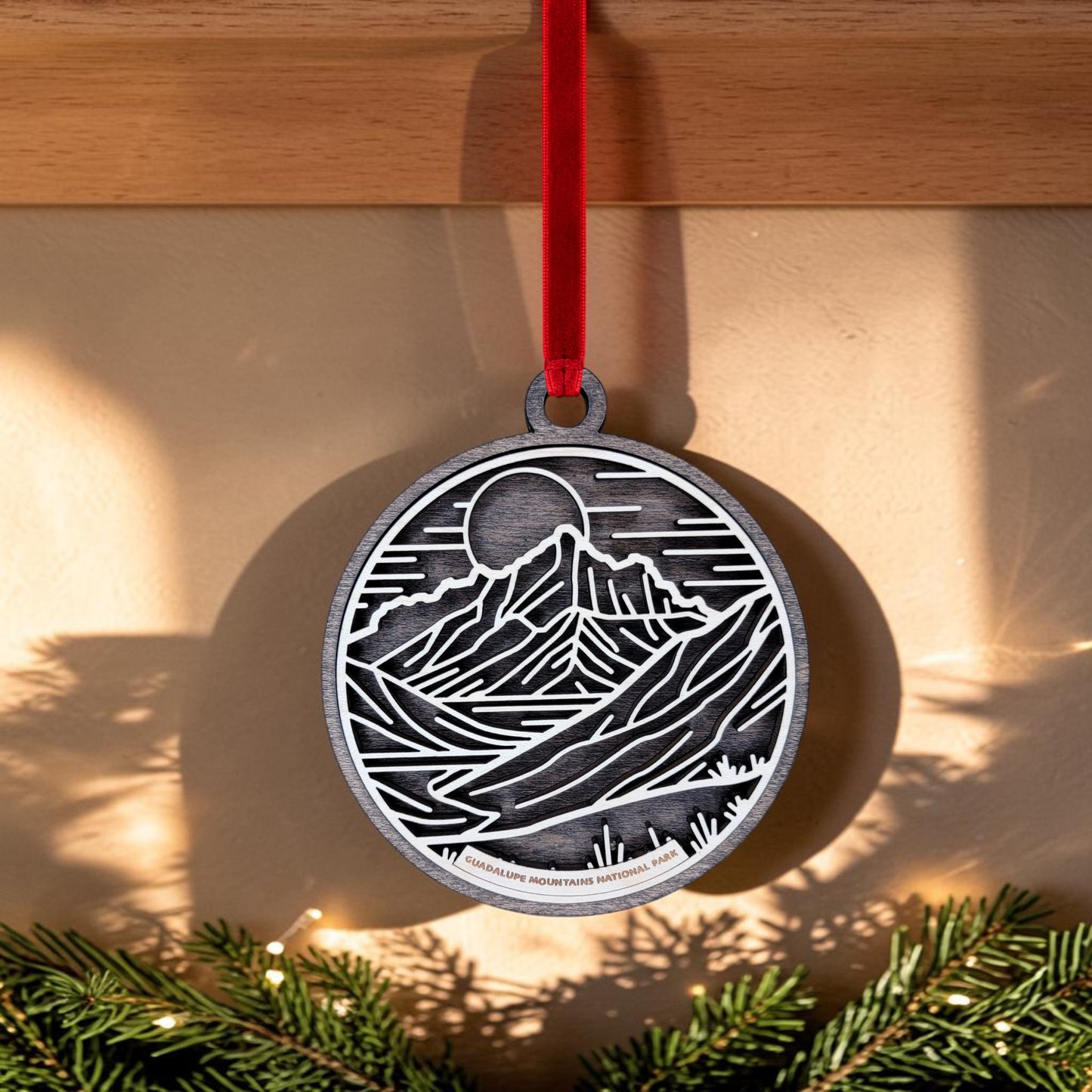 The Guadalupe Mountains National Park Line Art Christmas Ornament by Mirabilia Boutique, featuring a round metal design with a mountain and sun motif, hangs from a red ribbon in front of a beige wall. Pine branches and holiday lights decorate the bottom of the image.