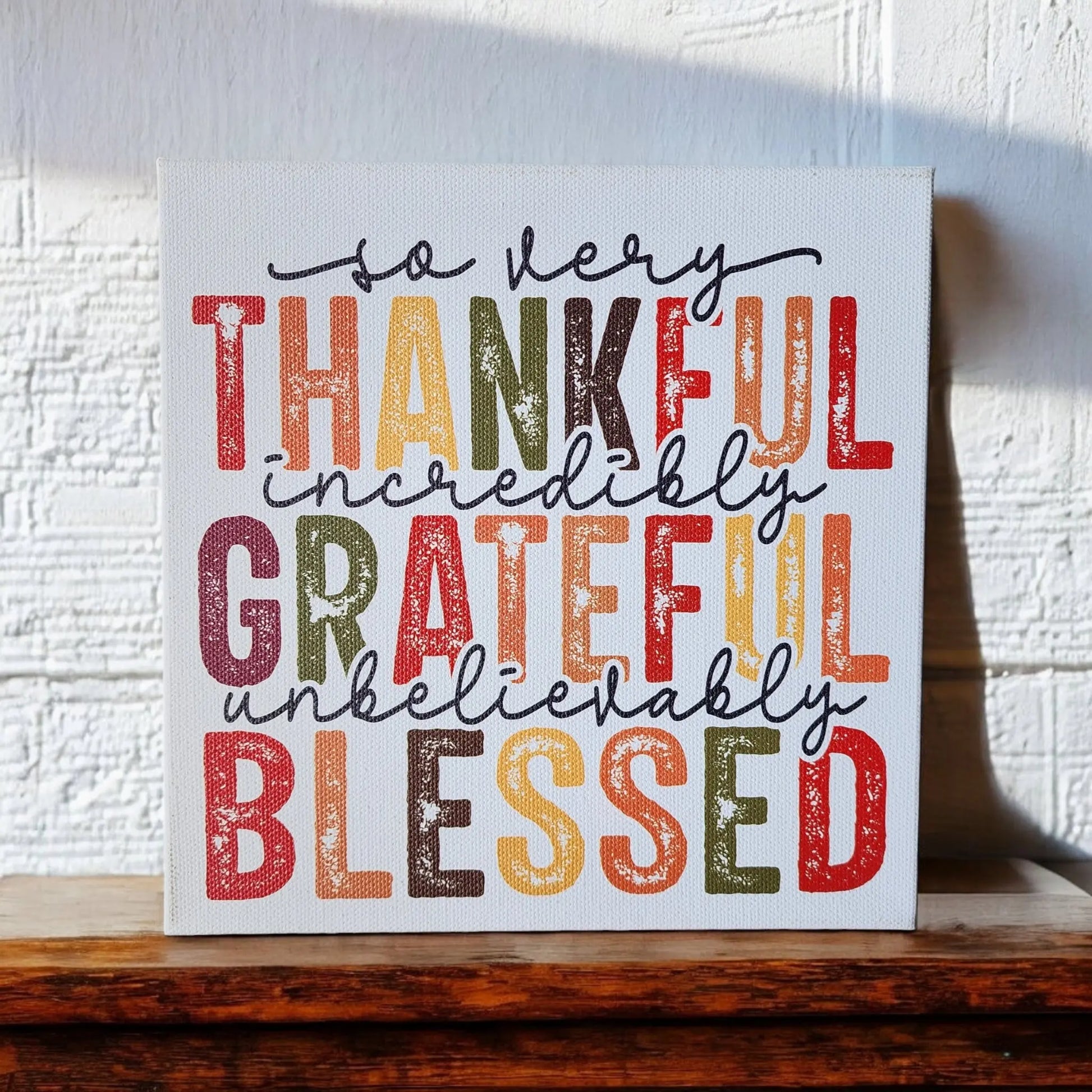A canvas from Mirabilia Boutique, featuring colorful and bold text, is propped against a white textured wall. The "Thankful Grateful Blessed Inspirational Sign for Seasonal Home Decor" reads: "so very Thankful, incredibly Grateful, unbelievably Blessed." Embracing Thanksgiving colors—shades of red, orange, yellow, green, and brown—this home decor sign is perfect for any cozy space. The canvas rests gracefully on a wooden surface.