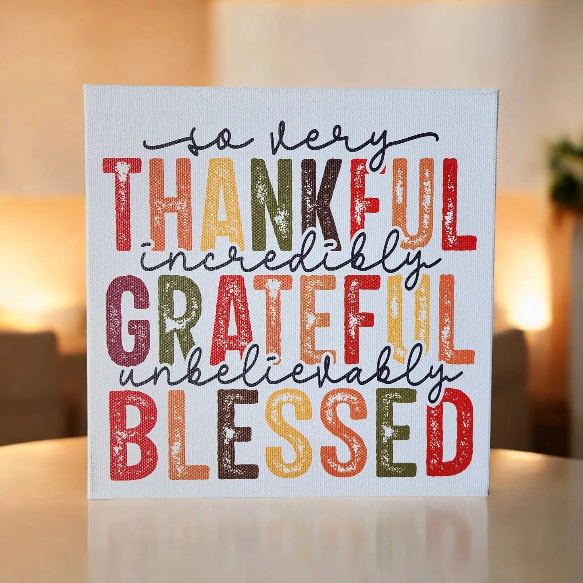 A square canvas from Mirabilia Boutique, titled "Thankful Grateful Blessed Inspirational Sign for Seasonal Home Decor," displays rustic, multicolored text that reads "So very thankful, incredibly grateful, unbelievably blessed." The background is artistically blurred, showcasing Thanksgiving colors in warm lighting with neutral tones and a subtle hint of greenery.