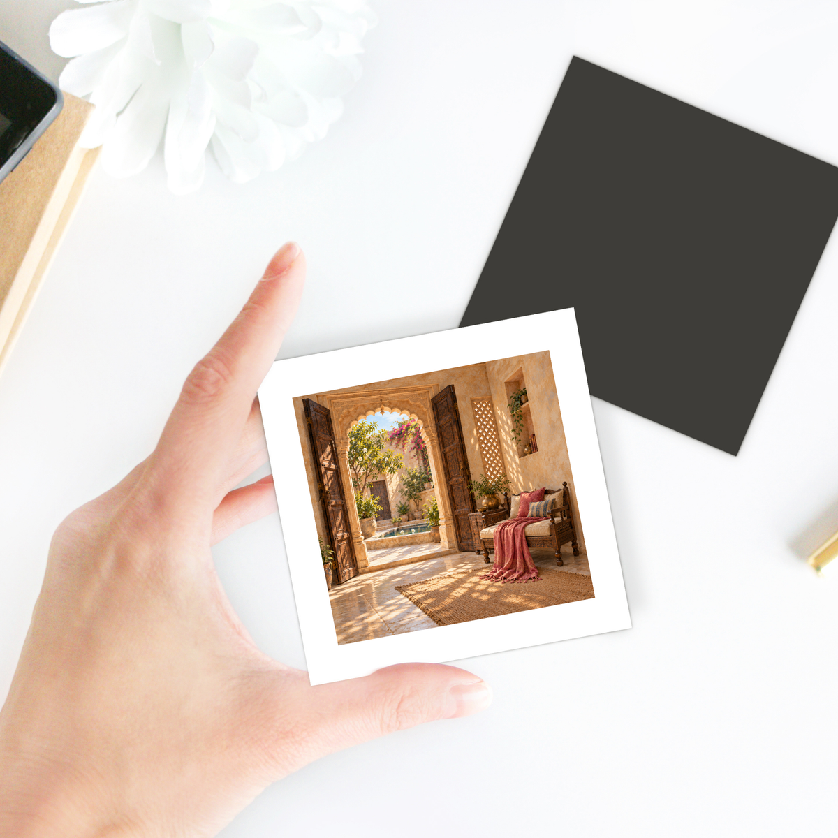 A hand holds a square The Door Left Open Magnet | Sand & Saffron decorative hardboard magnet featuring a photographic print of an open stone archway leading to a sunlit courtyard with a bench.