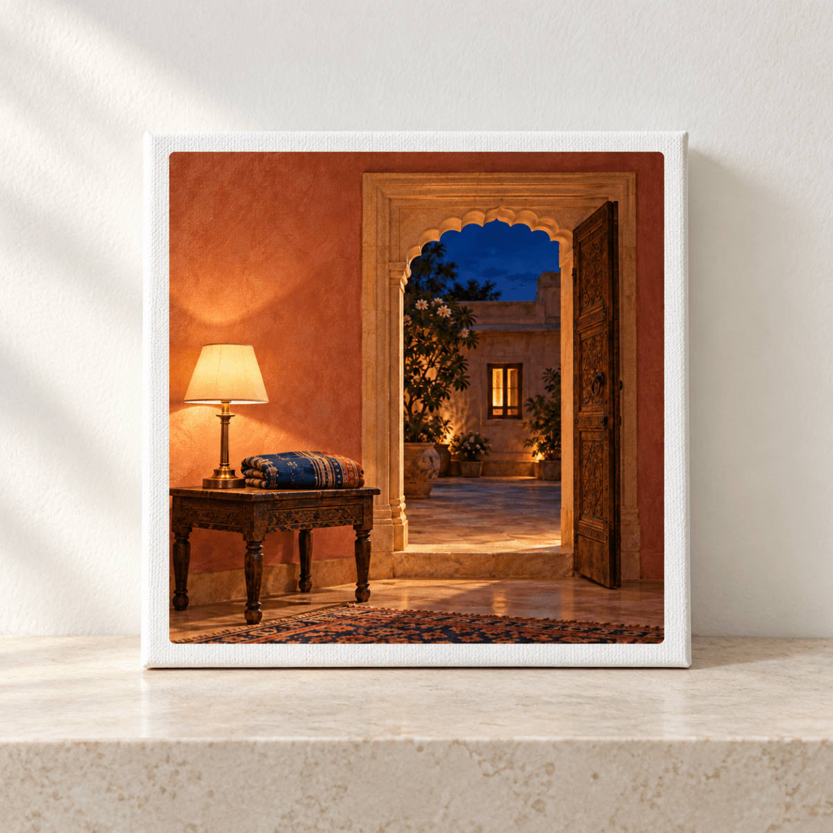 Square gallery-wrapped canvas titled The Light Still On Canvas | Sand & Saffron, depicting a wooden side table with a lamp and folded cloth next to an open doorway, revealing an evening courtyard art scene in terracotta tones.
