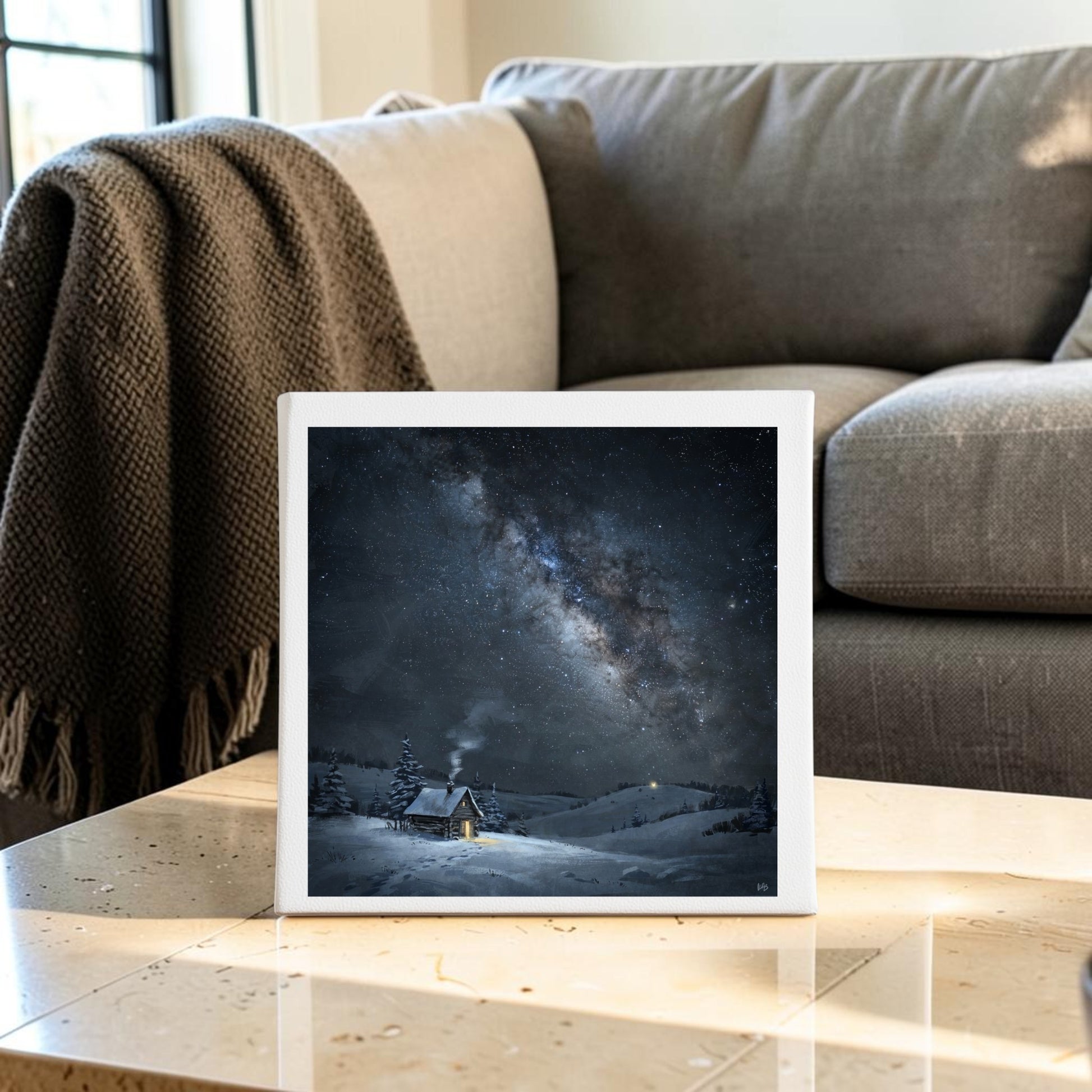 The Night Is Listening Milky Way Canvas Art (8x8) by Mirabilia Boutique, featuring a snowy cabin beneath a starry night sky, sits on a marble table in front of a gray couch and blanket.