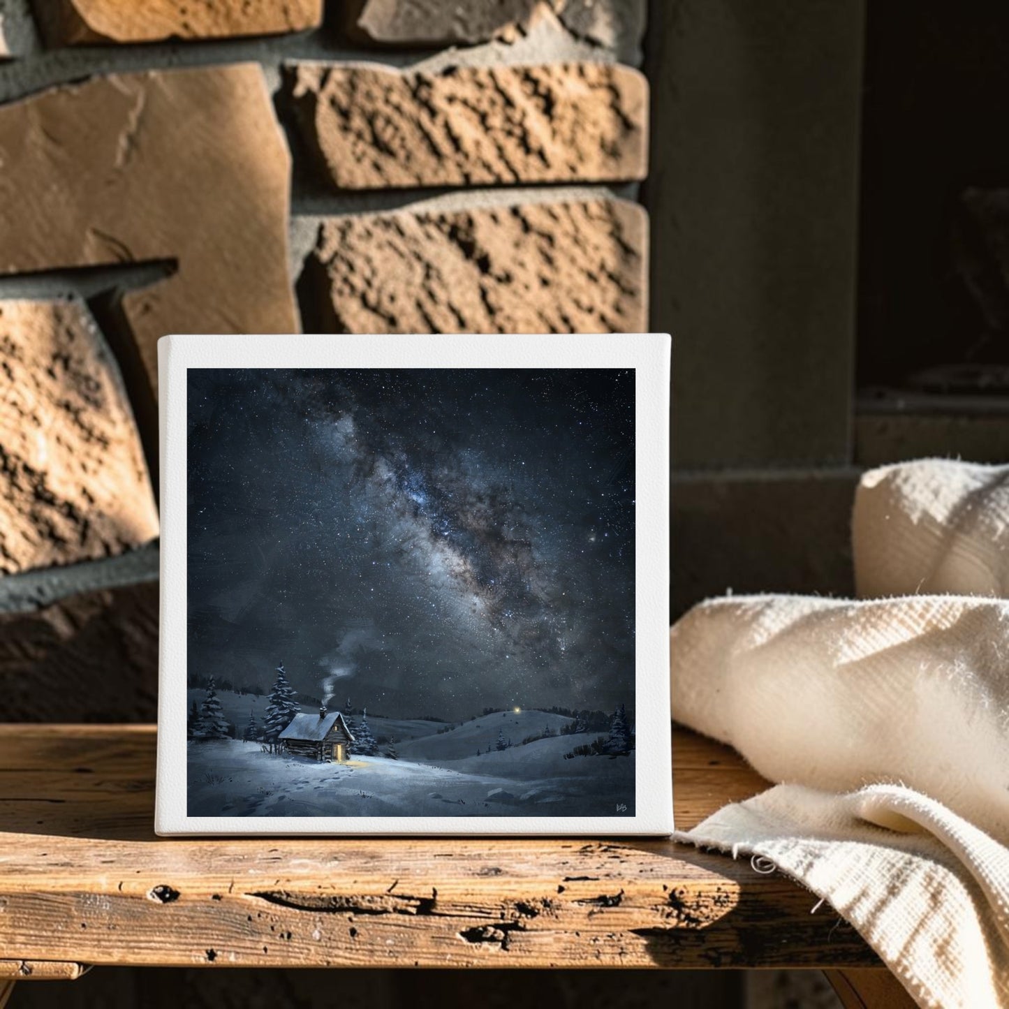 The Night Is Listening Milky Way Canvas Art by Mirabilia Boutique, in an 8x8 framed design, is displayed on a rustic wooden table with a white textured blanket beside it and a stone wall in the background. The artwork beautifully depicts a snowy cabin beneath a starry night sky.