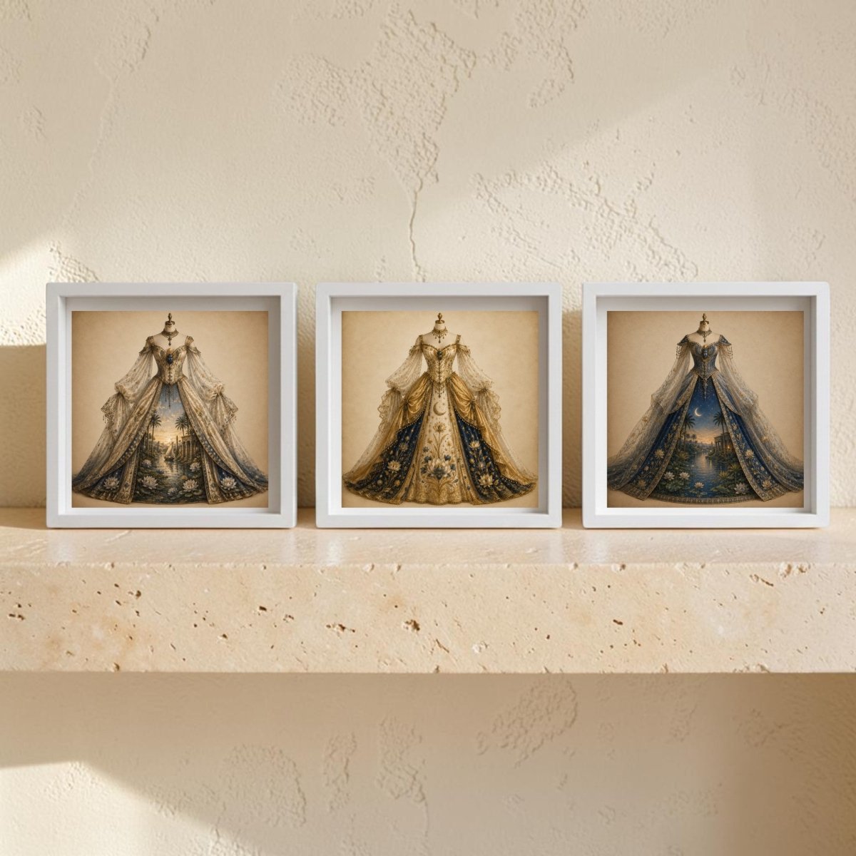 Three framed canvas prints from Mirabilia Boutique’s “The Nile Reverie | Curated Couture Artwork” collection, featuring ornate gowns in gold and blue hues, are displayed side by side on a beige stone shelf against a textured cream wall, creating an elegant curated couture arrangement.