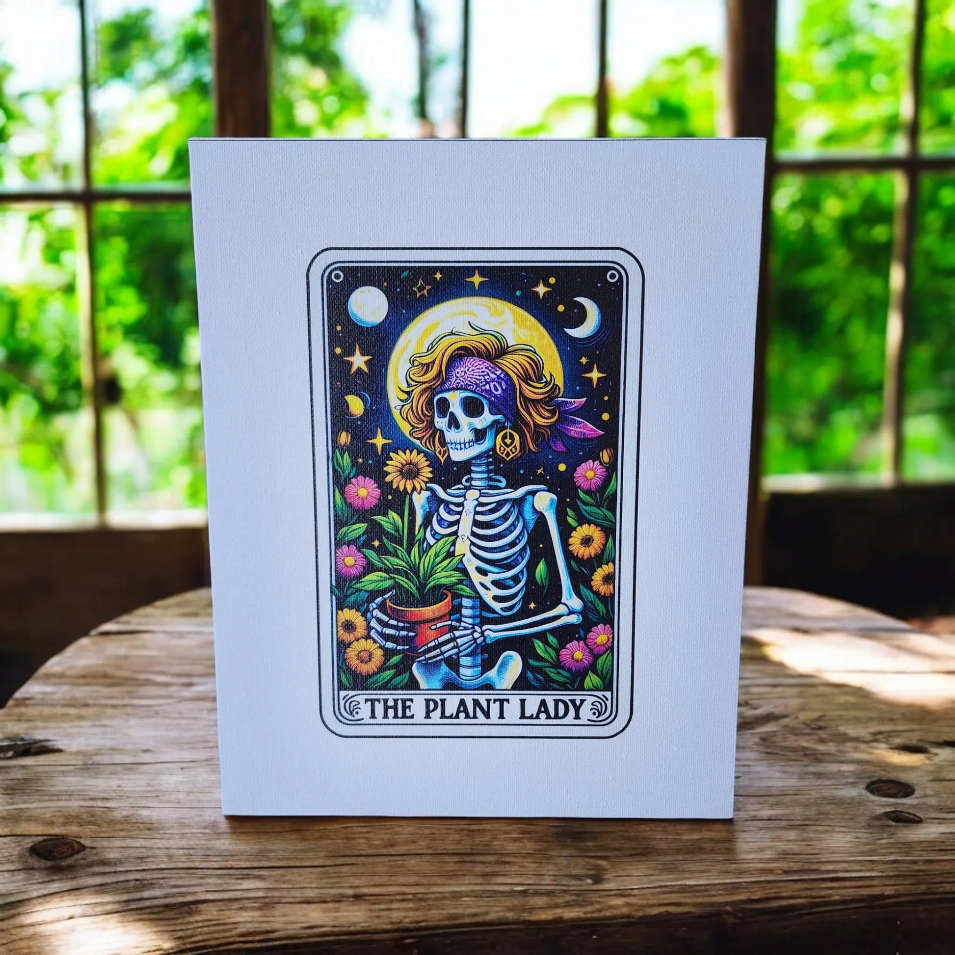 The Plant Lady Gardening 8x10 Canvas Sign by Mirabilia Boutique showcases a whimsical skeleton adorned in a hat and purple scarf, holding a potted plant amidst stars, sunflowers, and celestial symbols. Propped on a wooden table with bright green foliage visible through the window, this enchanting piece is perfect for tarot lovers.