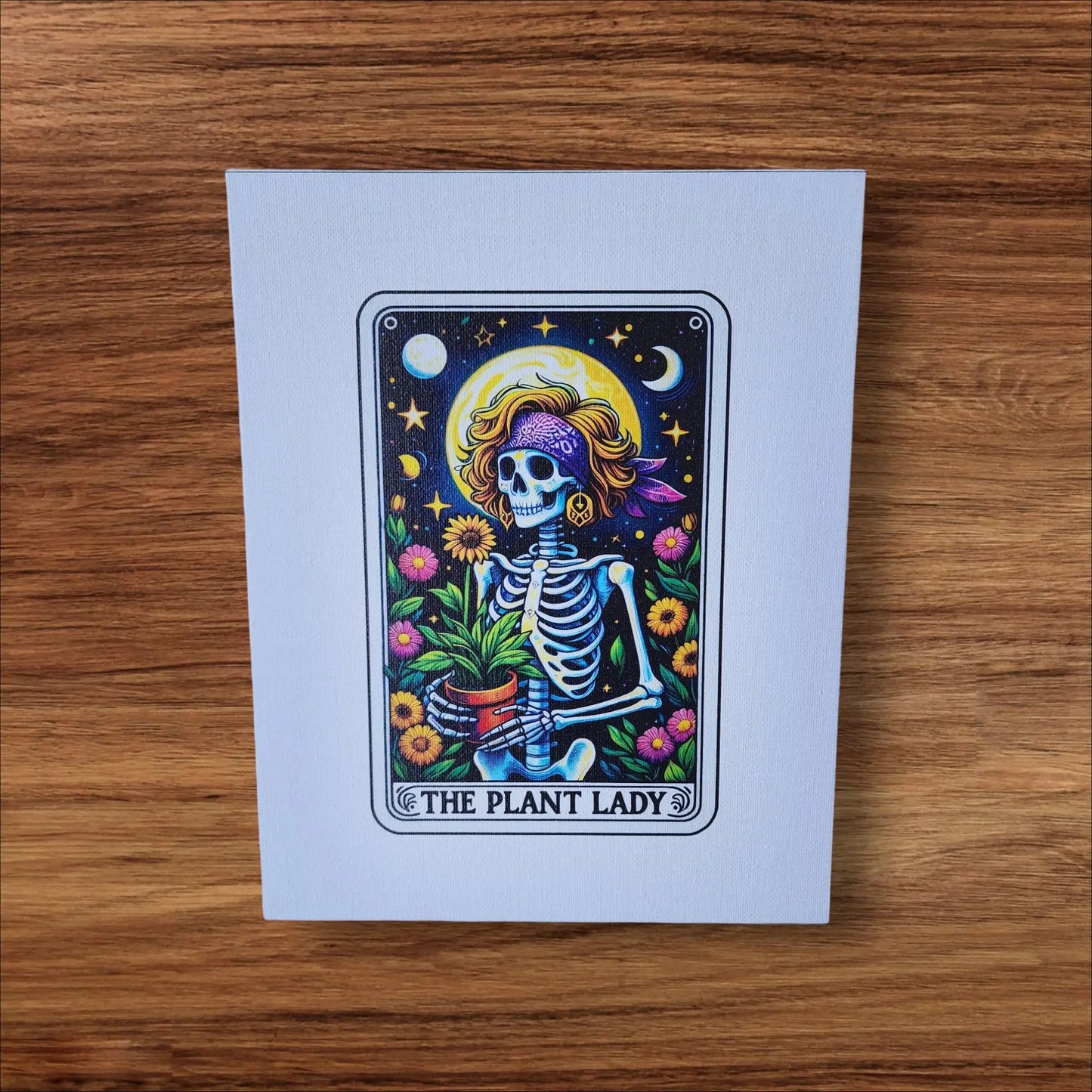The "The Plant Lady Gardening" 8x10 Canvas Sign by Mirabilia Boutique features a whimsical skeleton with a floral bandana, holding a potted plant amidst vibrant flowers, stars, and a crescent moon. Framed like a tarot card, this rectangular canvas is perfect for tarot lovers and adds charm to any space.