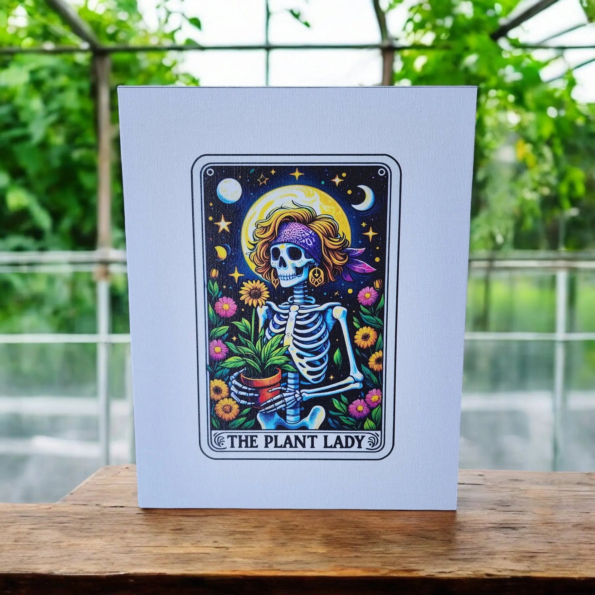 A colorful artwork titled "The Plant Lady Gardening 8x10 Canvas Sign" from Mirabilia Boutique depicts a whimsical skeleton wearing a headscarf, sunglasses, and holding a potted plant. Surrounded by flowers and celestial elements, the piece is displayed in a frame against a lush greenhouse backdrop—an enchanting sight for tarot lovers.