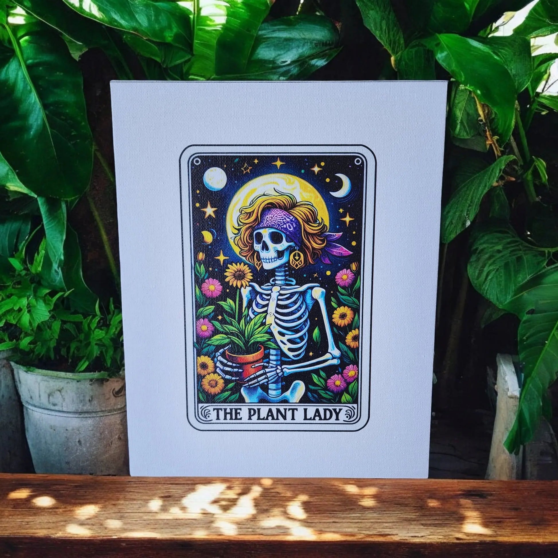 A colorful artwork from Mirabilia Boutique depicts a whimsical skeleton wearing a purple bandana and sunglasses, holding a potted plant surrounded by vibrant flowers, stars, and a crescent moon. The text at the bottom reads "The Plant Lady Gardening 8x10 Canvas Sign." The artwork is displayed outdoors with lush greenery as a backdrop.