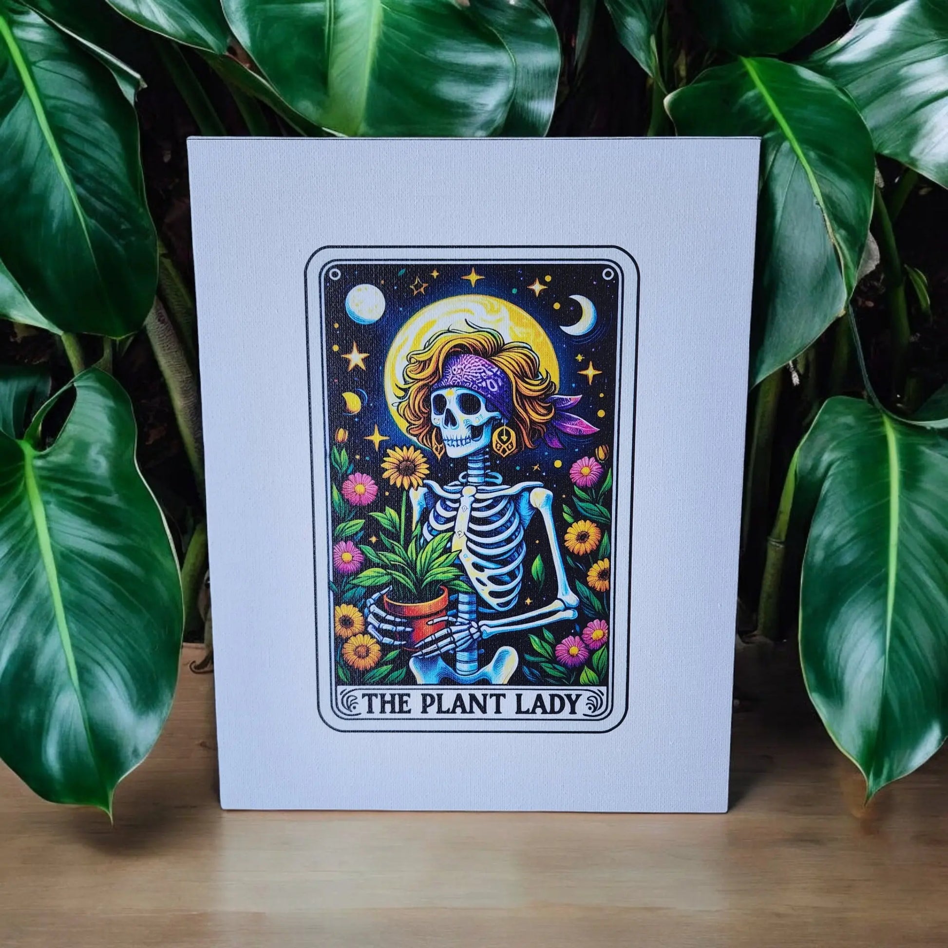 The "The Plant Lady Gardening 8x10 Canvas Sign" by Mirabilia Boutique features a colorful art print of a whimsical skeleton wearing a sun hat and purple bandana, holding a potted plant. The backdrop is adorned with stars, the moon, and flowers against lush green leaves, making it an ideal piece for tarot enthusiasts.