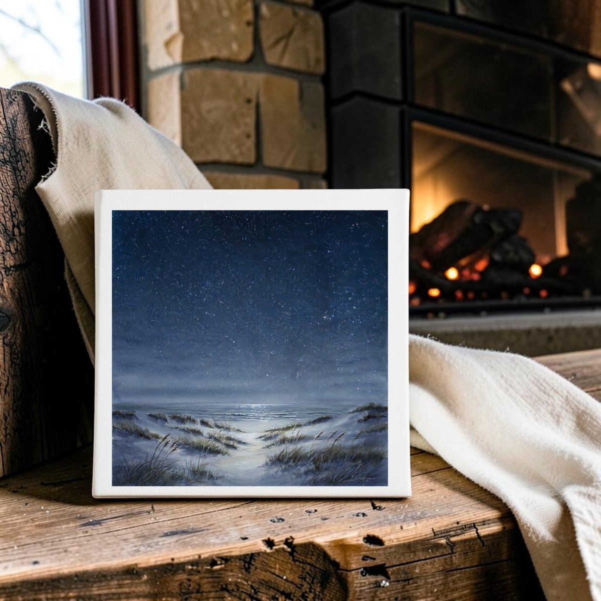 The Sea Keeps Its Own Stars 8x8 Canvas by Mirabilia Boutique, a serene coastal decor piece featuring a starry night sky over sand dunes, is displayed on a wooden bench near a fireplace with a cream-colored blanket draped nearby.