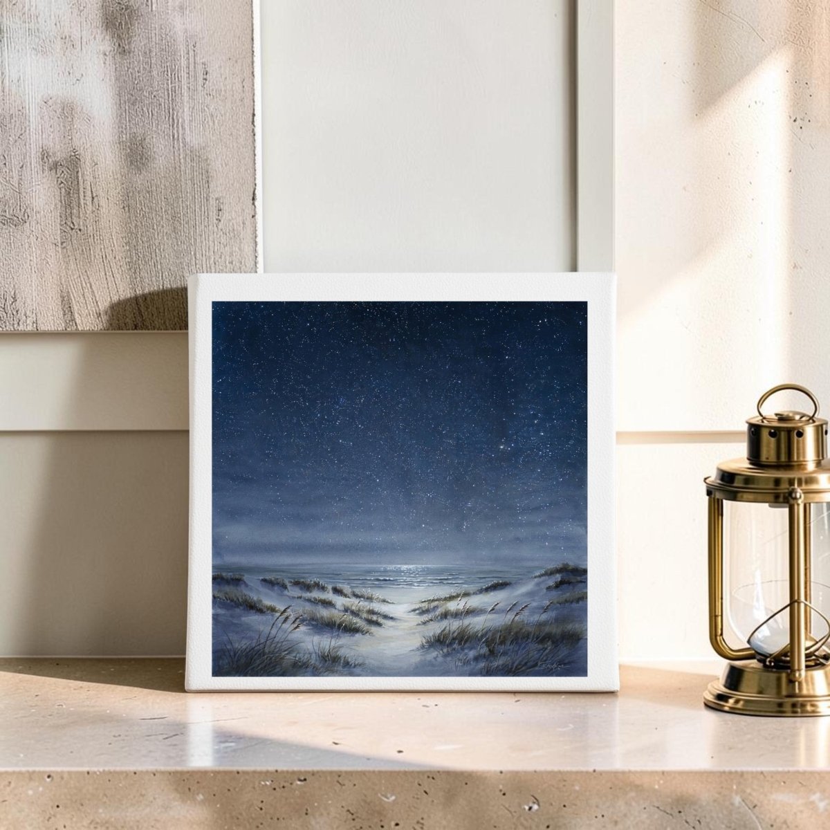 The Sea Keeps Its Own Stars 8x8 Canvas by Mirabilia Boutique, featuring a starry night sky over sandy dunes with grass, sits on a beige mantel beside a brass lantern—perfect for adding serene coastal decor to your space.