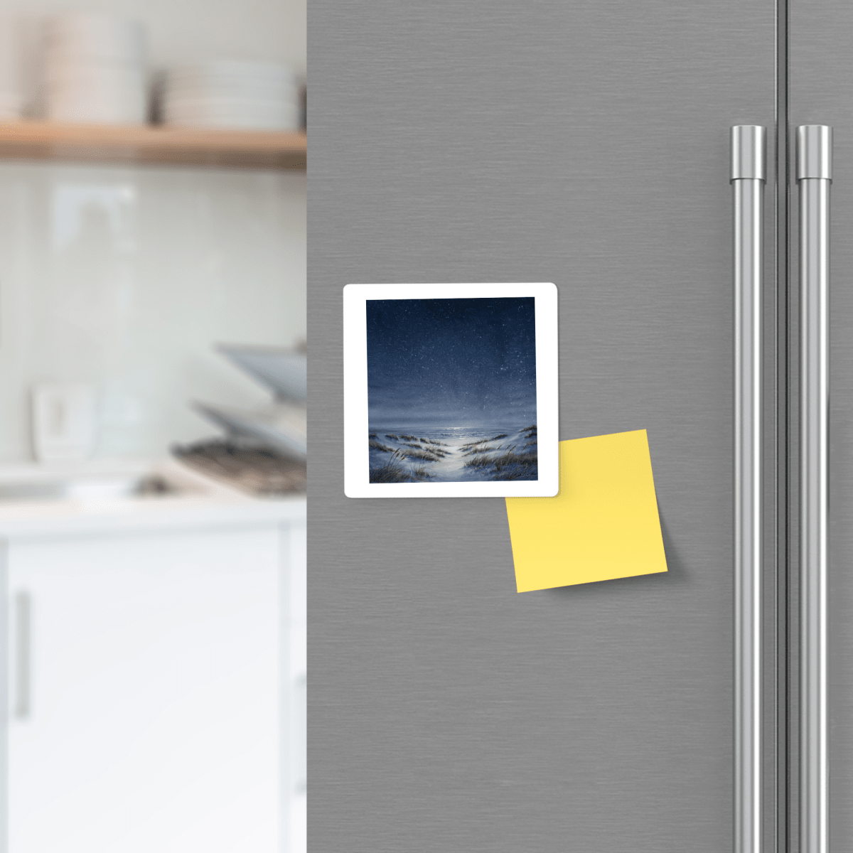 A refrigerator door featuring a photo of a snowy night scene, a blank yellow sticky note attached, and Mirabilia Boutique’s The Sea Keeps Its Own Stars Magnet placed beside it. Blurred kitchen shelves with dishes appear in the background.