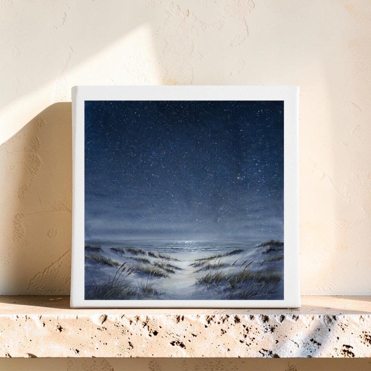 The Sea Keeps Its Own Stars 8x8 Canvas by Mirabilia Boutique features a tranquil moonlit beach scene with sand dunes and grasses, displayed on a textured stone shelf against a beige wall. The serene night sky is filled with stars that gently reflect on the water, making this piece a stunning example of starry night wall art.