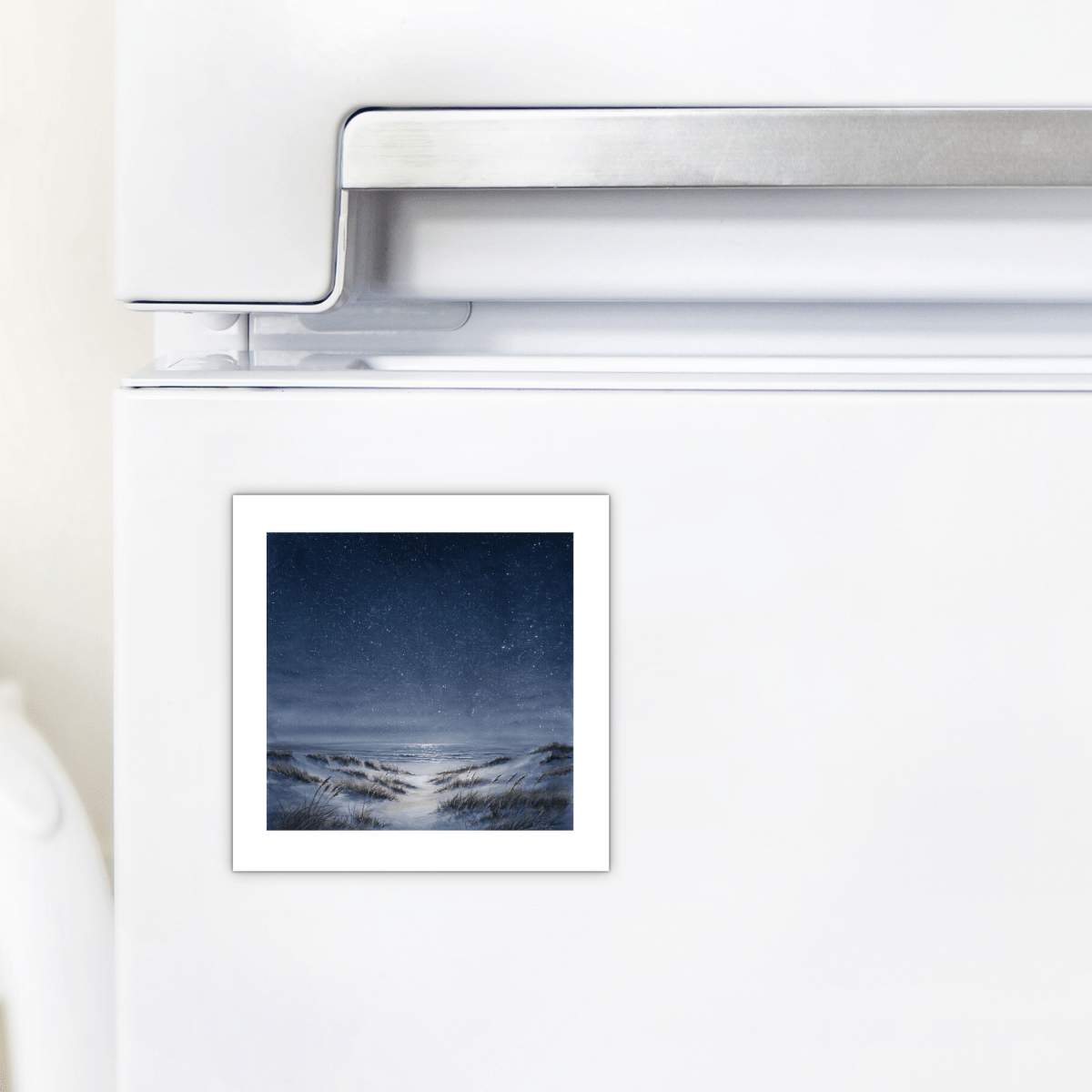 A white refrigerator features The Sea Keeps Its Own Stars Magnet by Mirabilia Boutique—a small, square magnet depicting a snowy night landscape beneath a starry sky—placed near the handle. This charming piece brings coastal serenity and a moonlit ambiance to your kitchen décor, infusing your space with the tranquil beauty of a starlit shore.